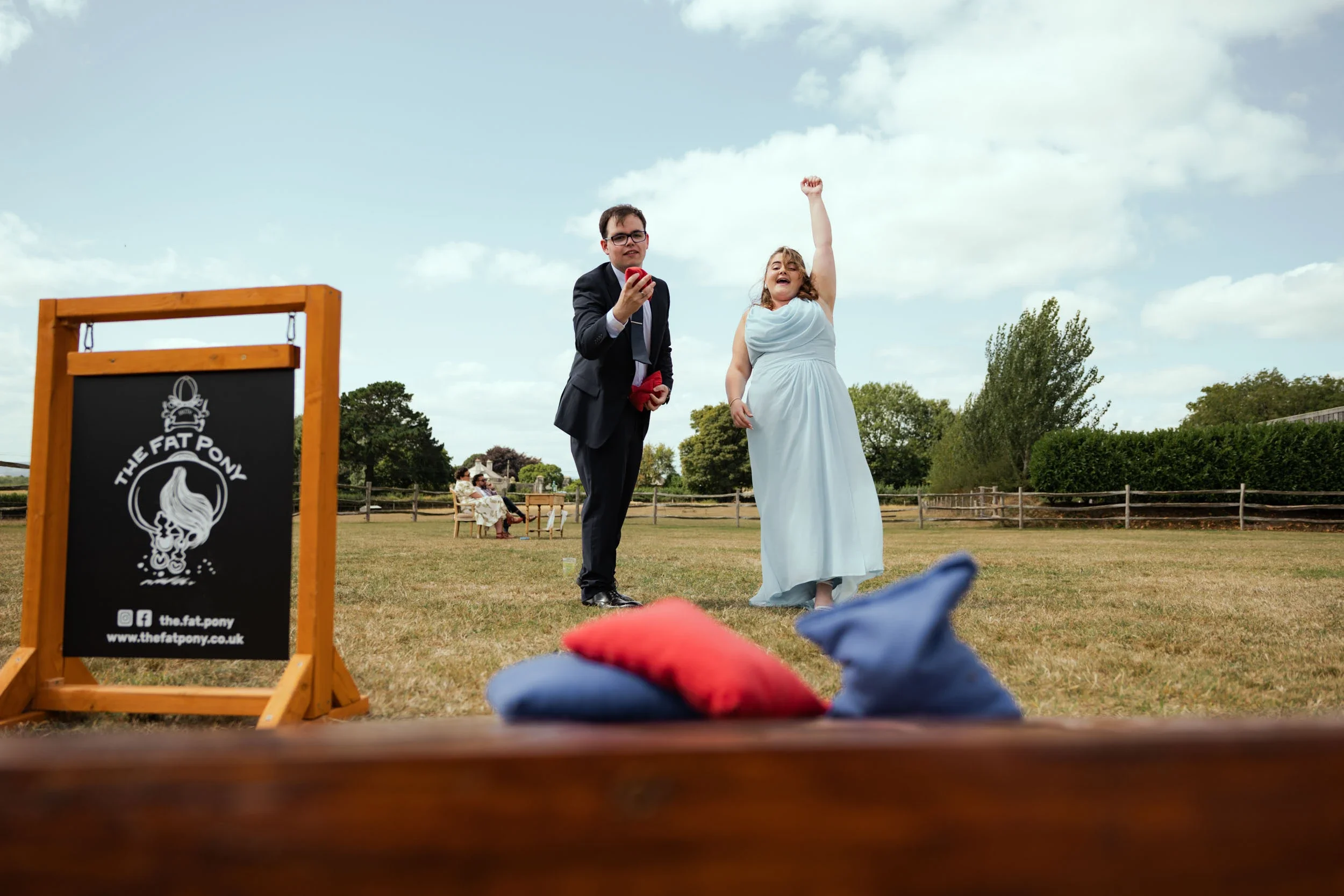 A bride in a white dress and a groom in a suit are at an outdoor wedding ceremony, with the bride celebrating and the groom holding a red object, on a grassy area with benches and trees in the background.