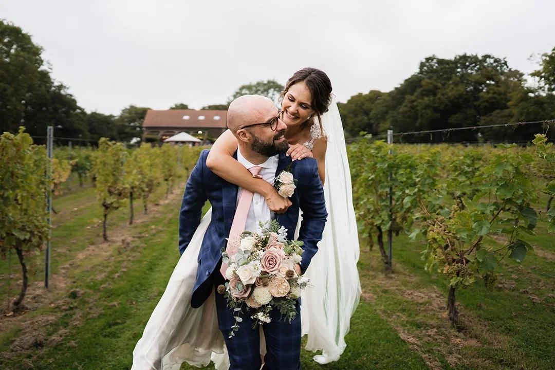 Bride and groom in a vineyard, with the bride on the groom's back, smiling, holding a bouquet of flowers.