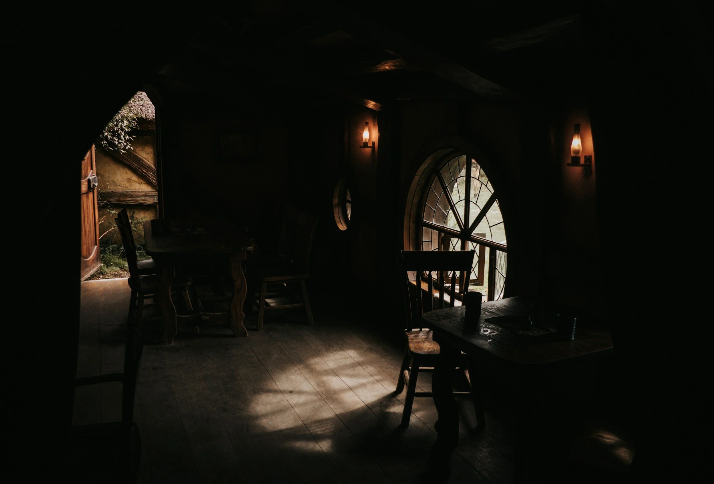 Dimly lit rustic interior with wooden furniture and a large arched window allowing natural light to enter