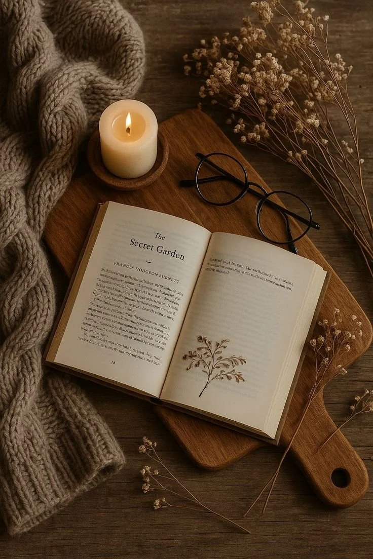 An open book titled 'The Secret Garden' resting on a wooden board, with a pair of black-rimmed glasses, a lit candle in a wooden holder, a cozy knitted blanket, and dried flowers on a wooden surface.
