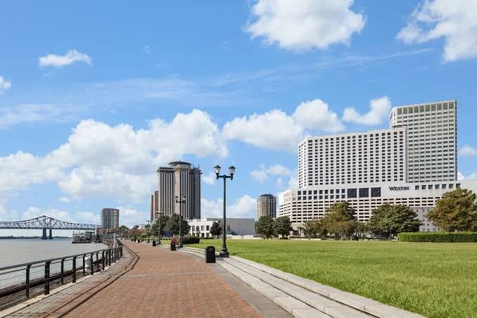 Scenic view of a city waterfront with a brick walkway, green grass, modern buildings, and a bridge over the river under a partly cloudy sky.