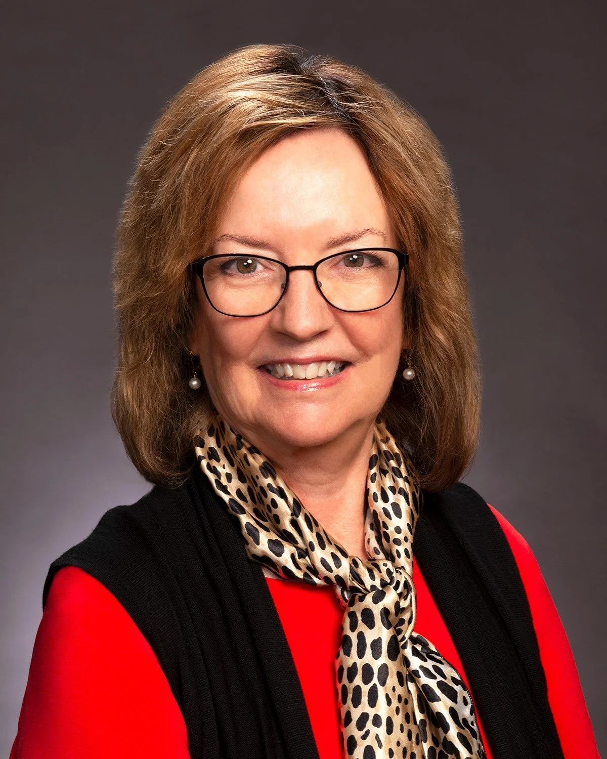 A woman with shoulder-length wavy red hair and blue eyes, smiling, wearing a tan blazer, against a plain white background.