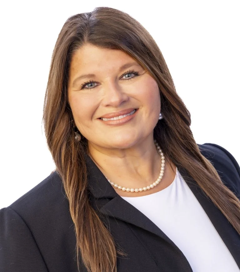 A woman with brown hair, wearing a dark blazer, white top, pearl necklace, and pearl earrings, smiling at the camera.