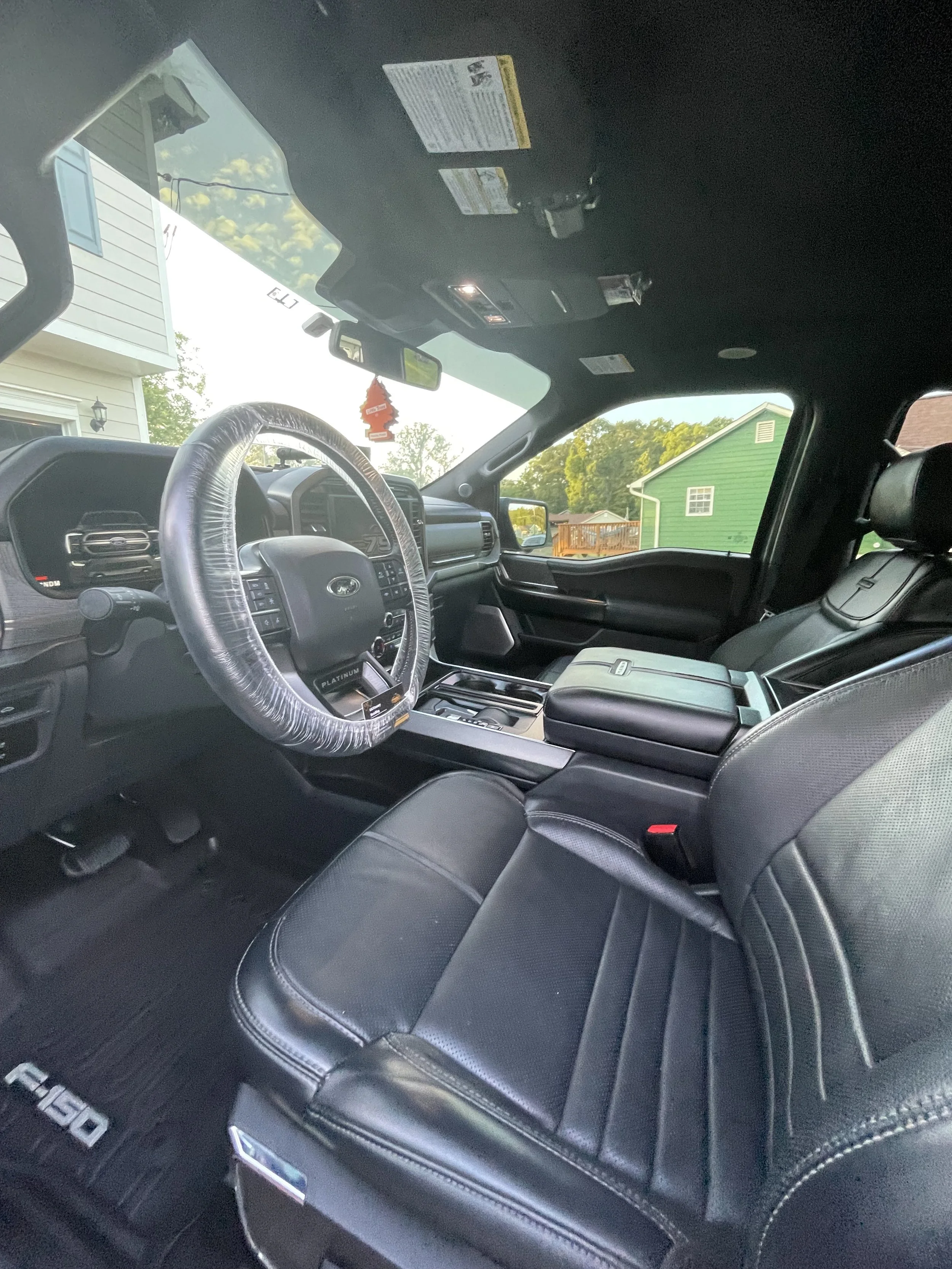 Interior of a black Ford F-150 truck showing the dashboard, steering wheel, and front seats. Outside the window, houses and trees are visible.