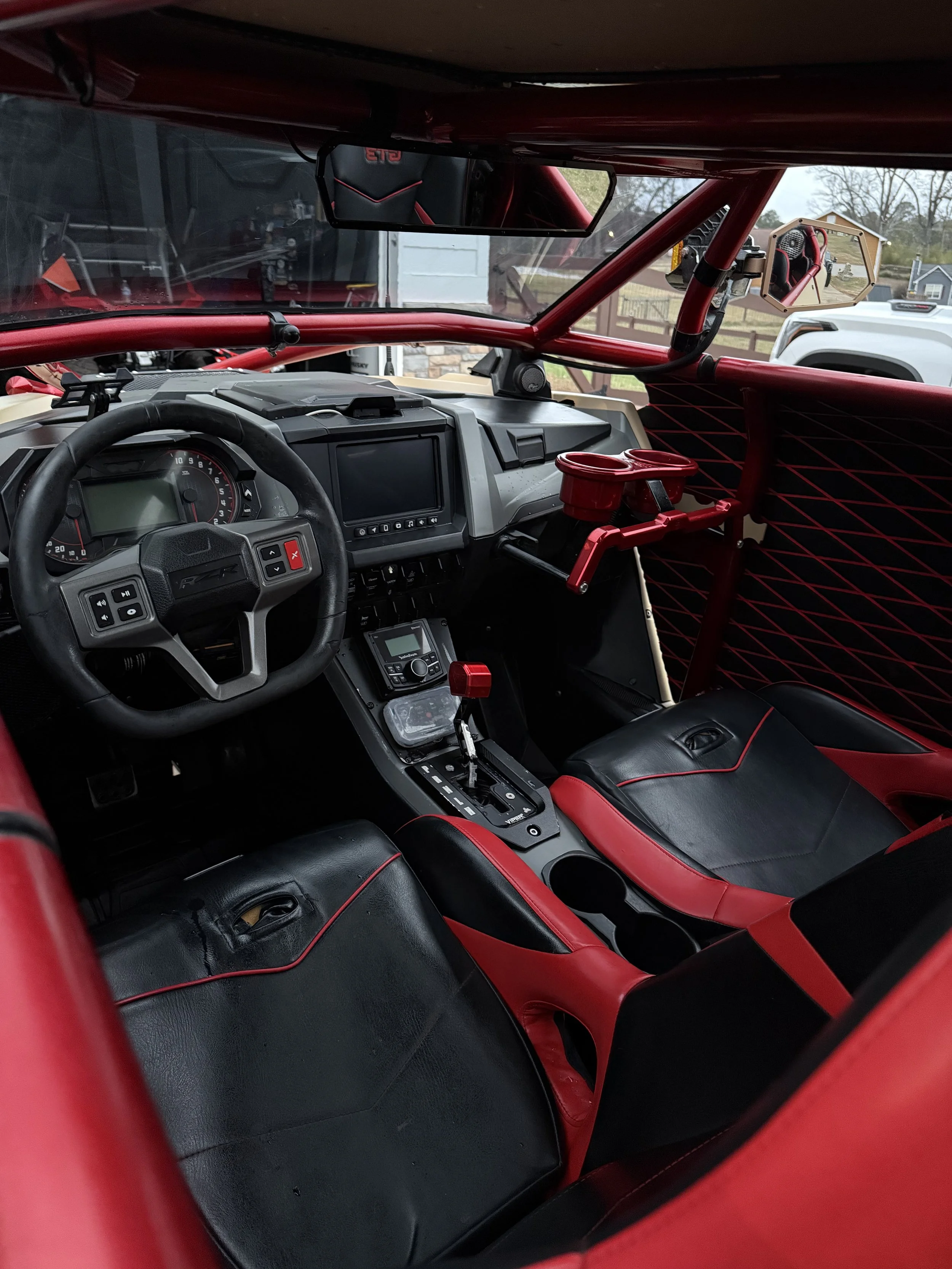 The interior of a race car showing black and red padded seats, a steering wheel, a gear shifter, a digital display, and a red cup holder. The dashboard has various controls and a display screen. The garage window and outdoor scenery are visible throu