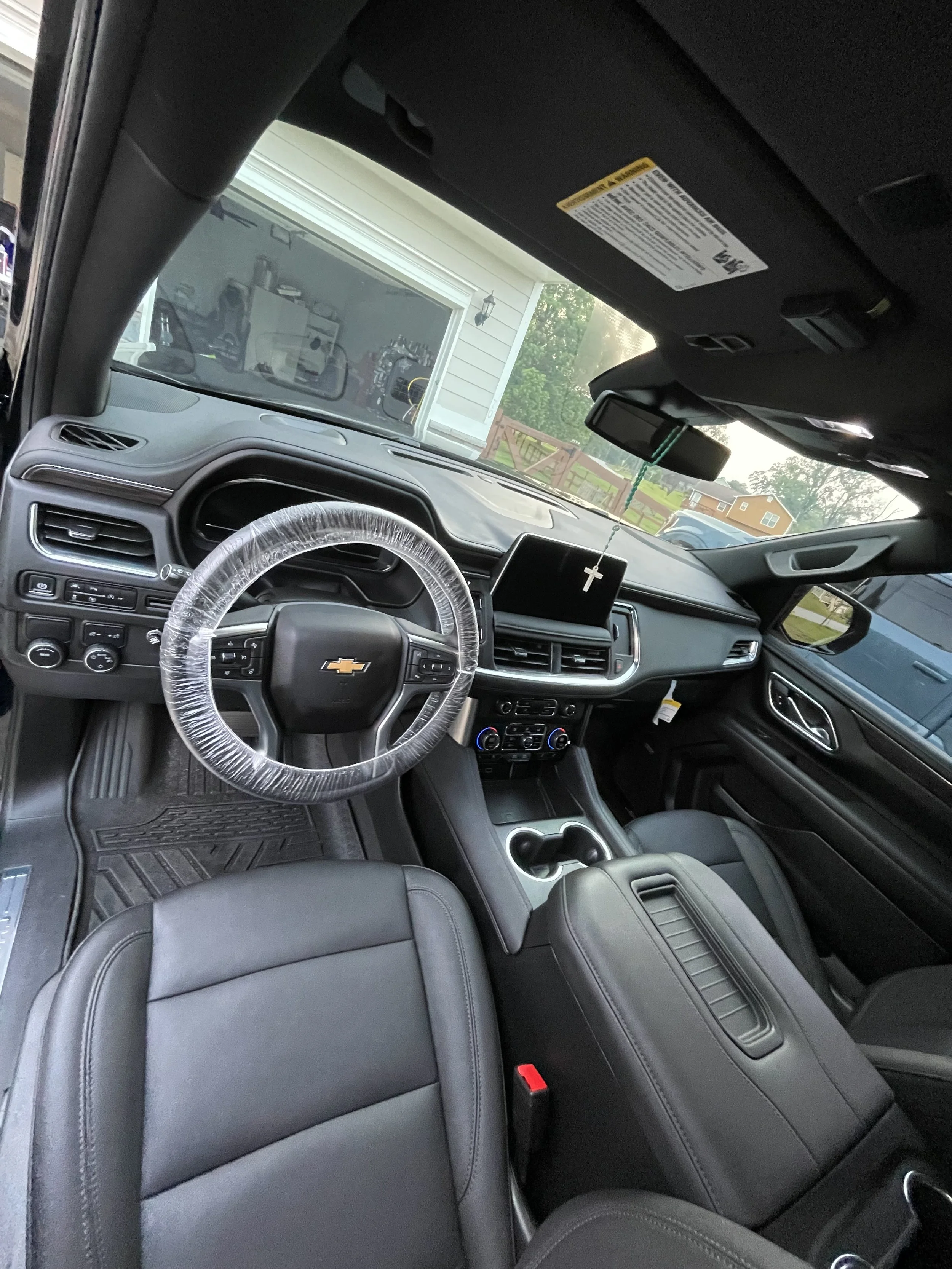 The interior of a Chevrolet vehicle showing the dashboard, steering wheel with transparent cover, center console, gear shift, and front seats. The dashboard has a display screen and control buttons, with a religious cross hanging from the rearview mi