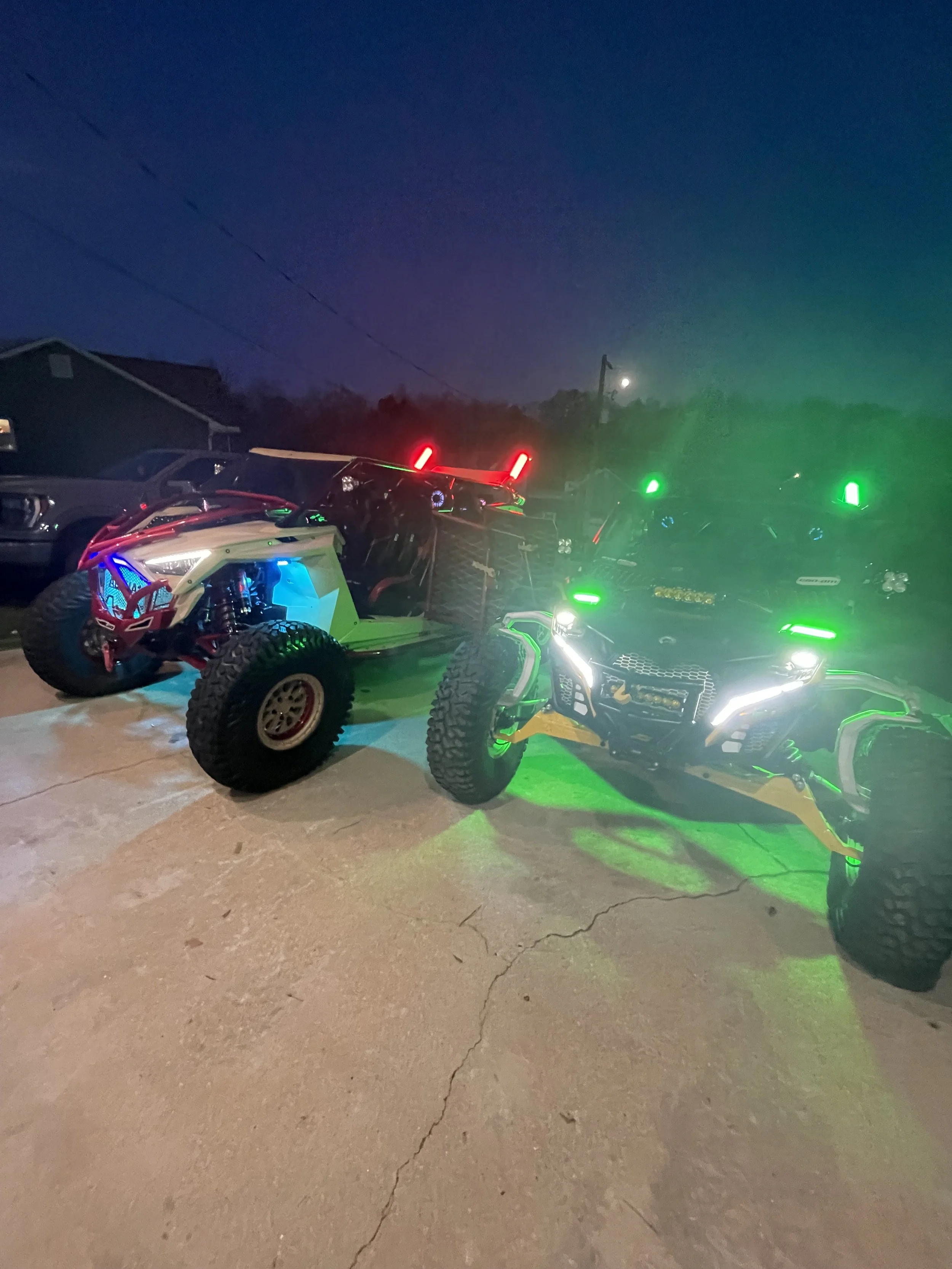 Two off-road vehicles with illuminated LED lights parked on a concrete surface during nighttime, with a dark sky and some trees and houses in the background.