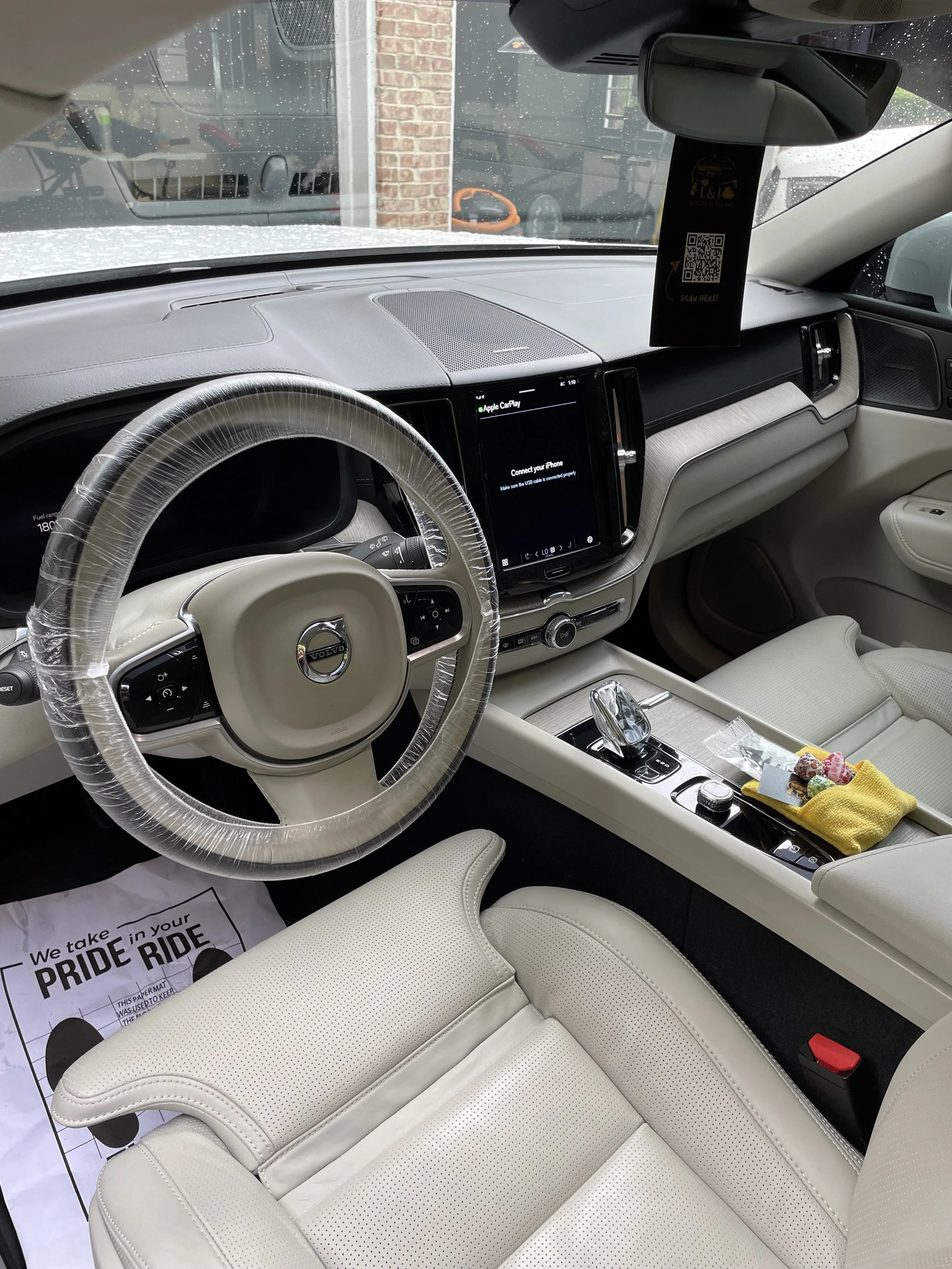 Interior of a luxury Volvo vehicle with beige leather seats, a modern dashboard featuring a large touchscreen display, a steering wheel with controls, and some items placed on the center console, including a yellow cloth and small objects.