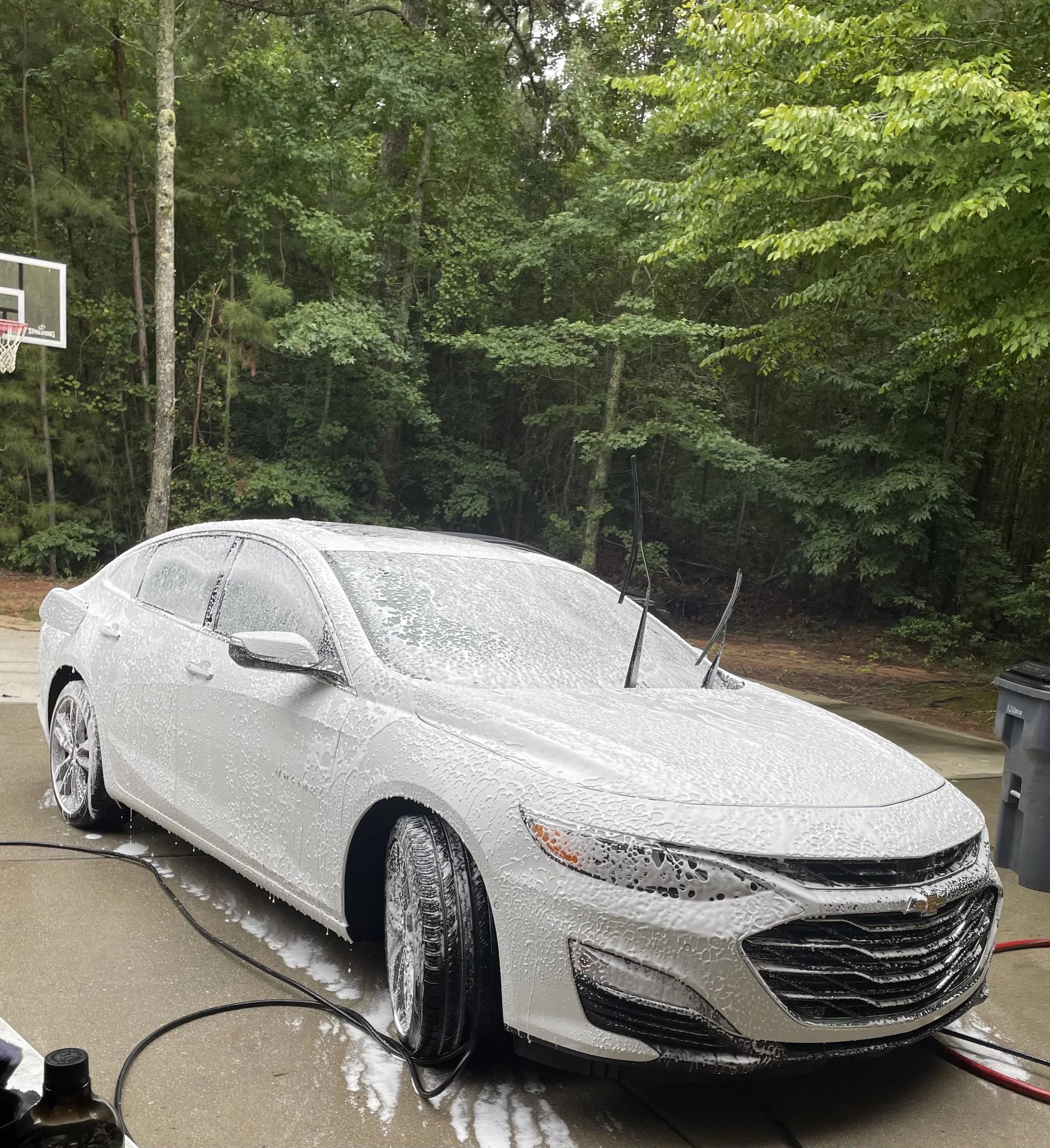 A white sedan is being washed with soap, with foam covering the car. The car is on a wet driveway, surrounded by green trees and a basketball hoop on the left.