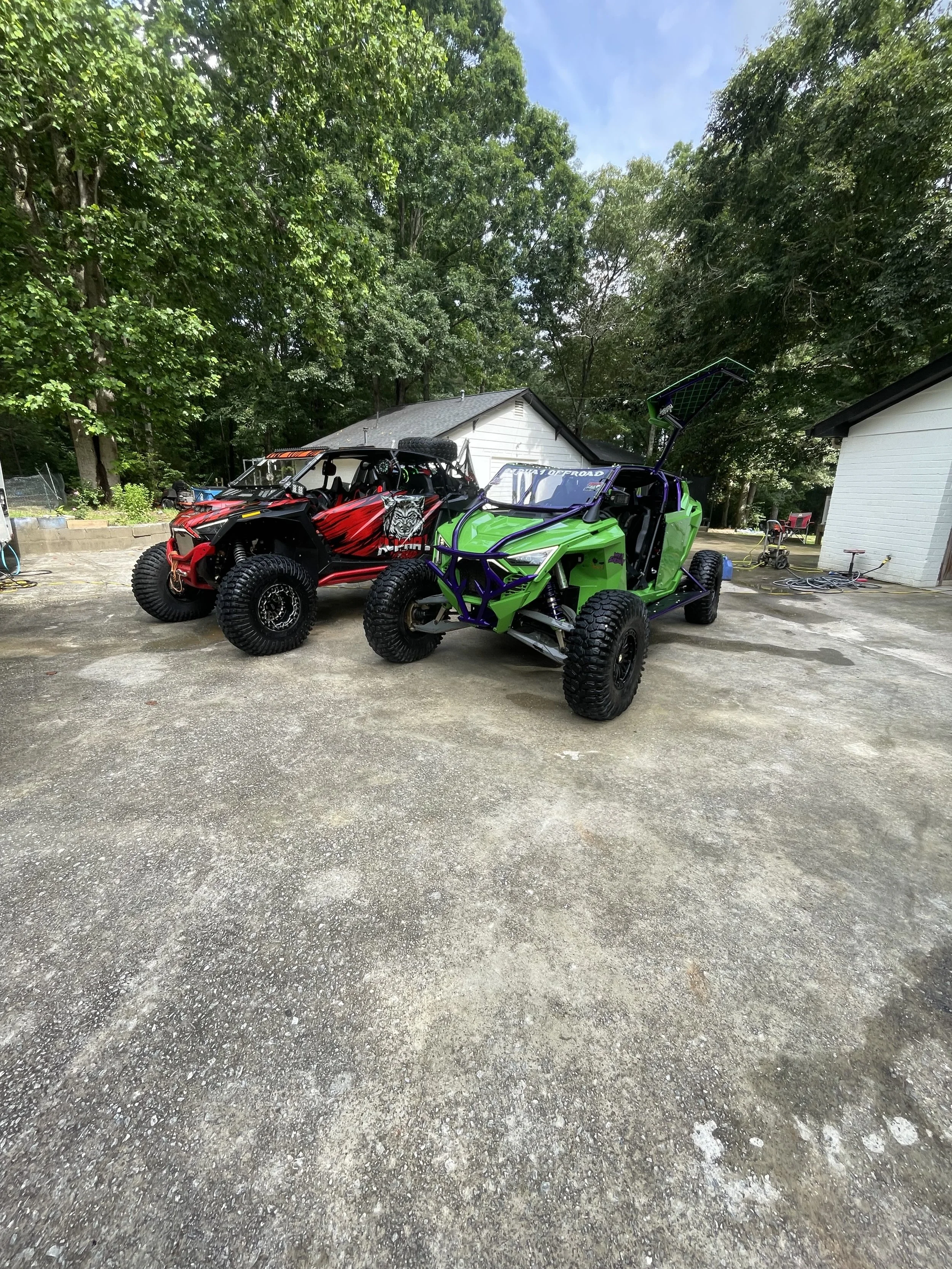 Two off-road vehicles parked on a concrete driveway, one red and black, and the other green and purple, with trees and buildings in the background.