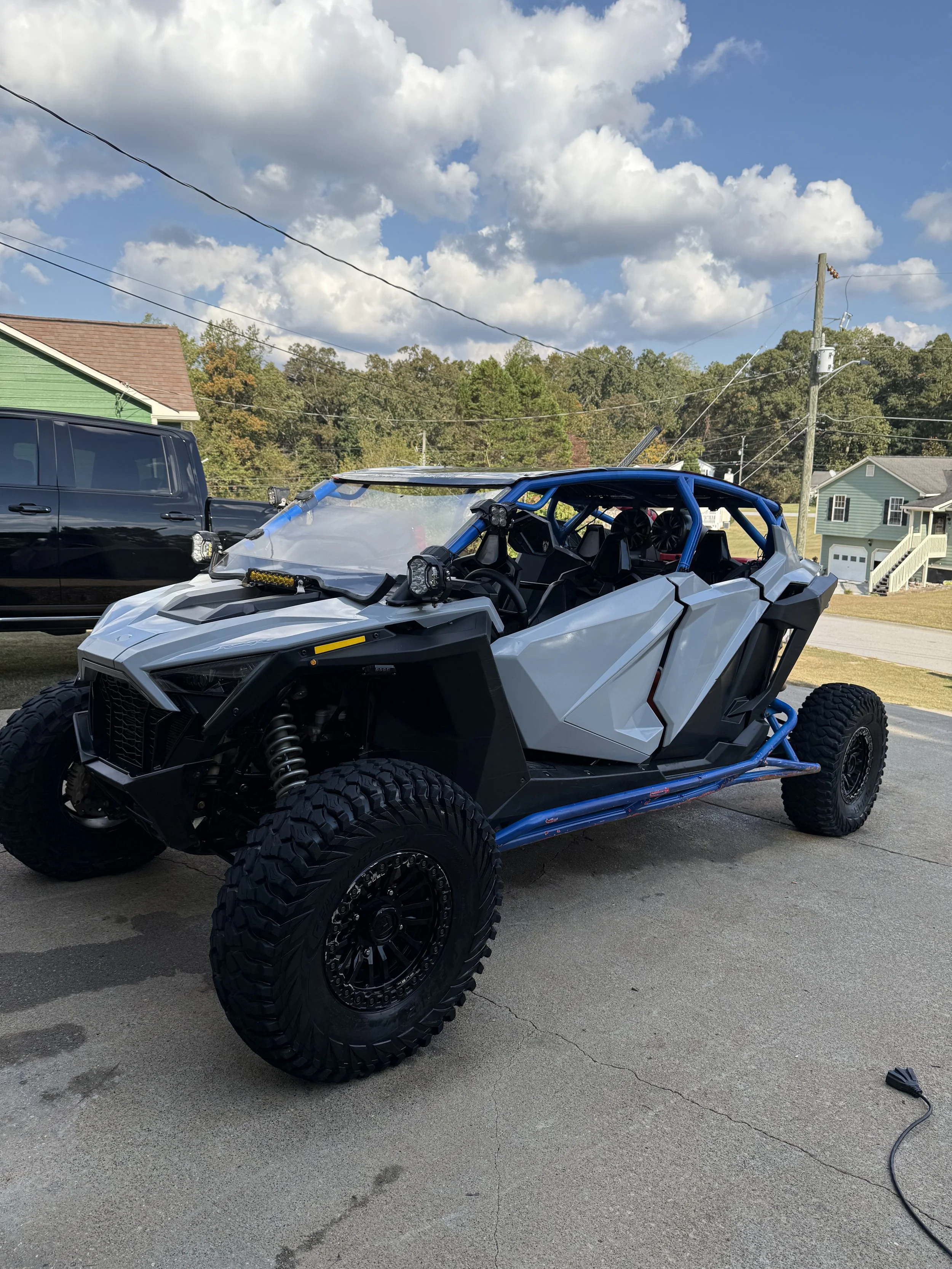 A white off-road vehicle with a blue roll cage and large tires parked on a driveway. There is a black pickup truck in the background, along with houses and a partly cloudy sky.