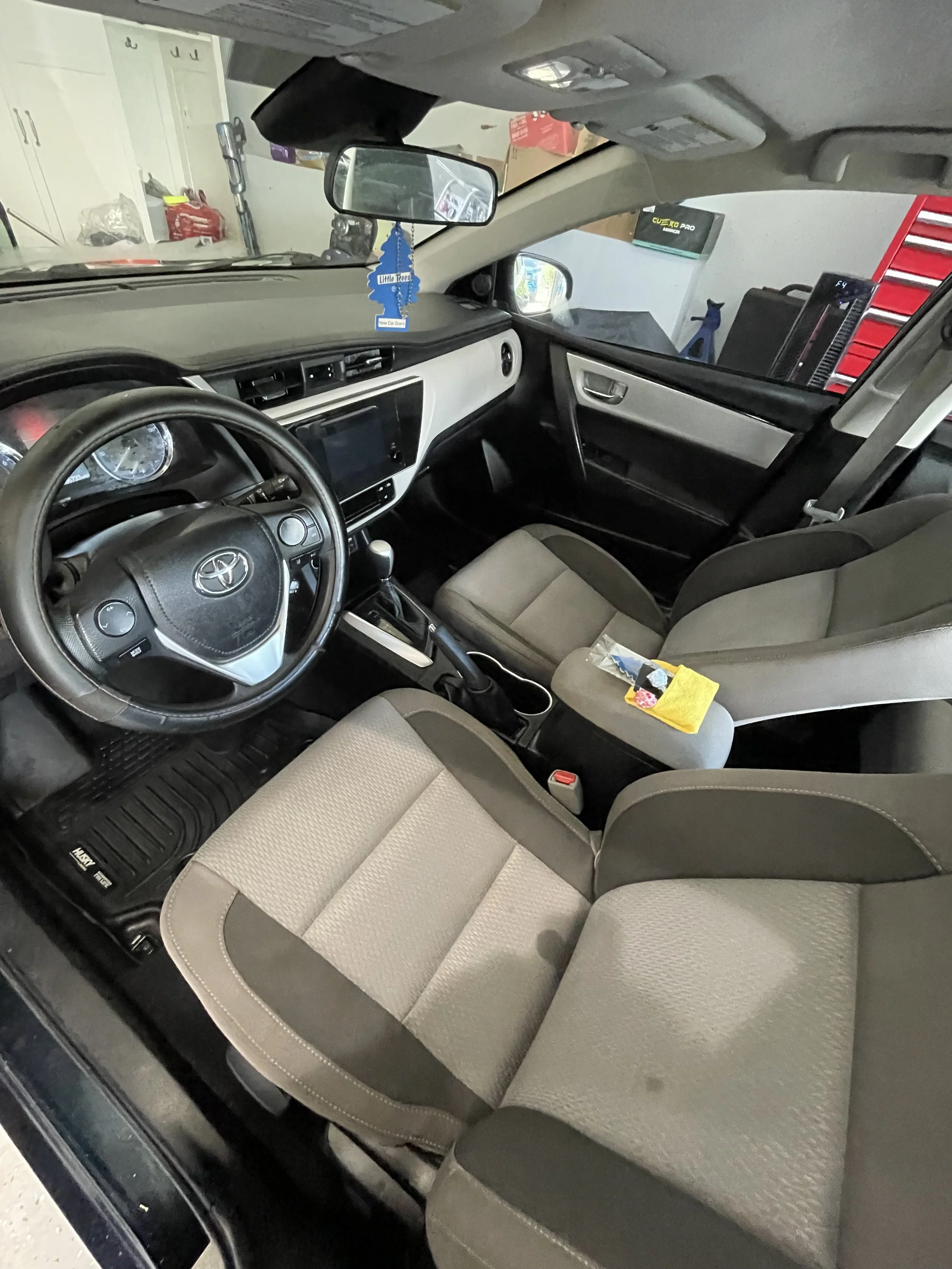 The interior of a Toyota vehicle showing the front seats, steering wheel, dashboard, and rearview mirror. The dashboard has a touchscreen display, and there are objects hanging from the rearview mirror.