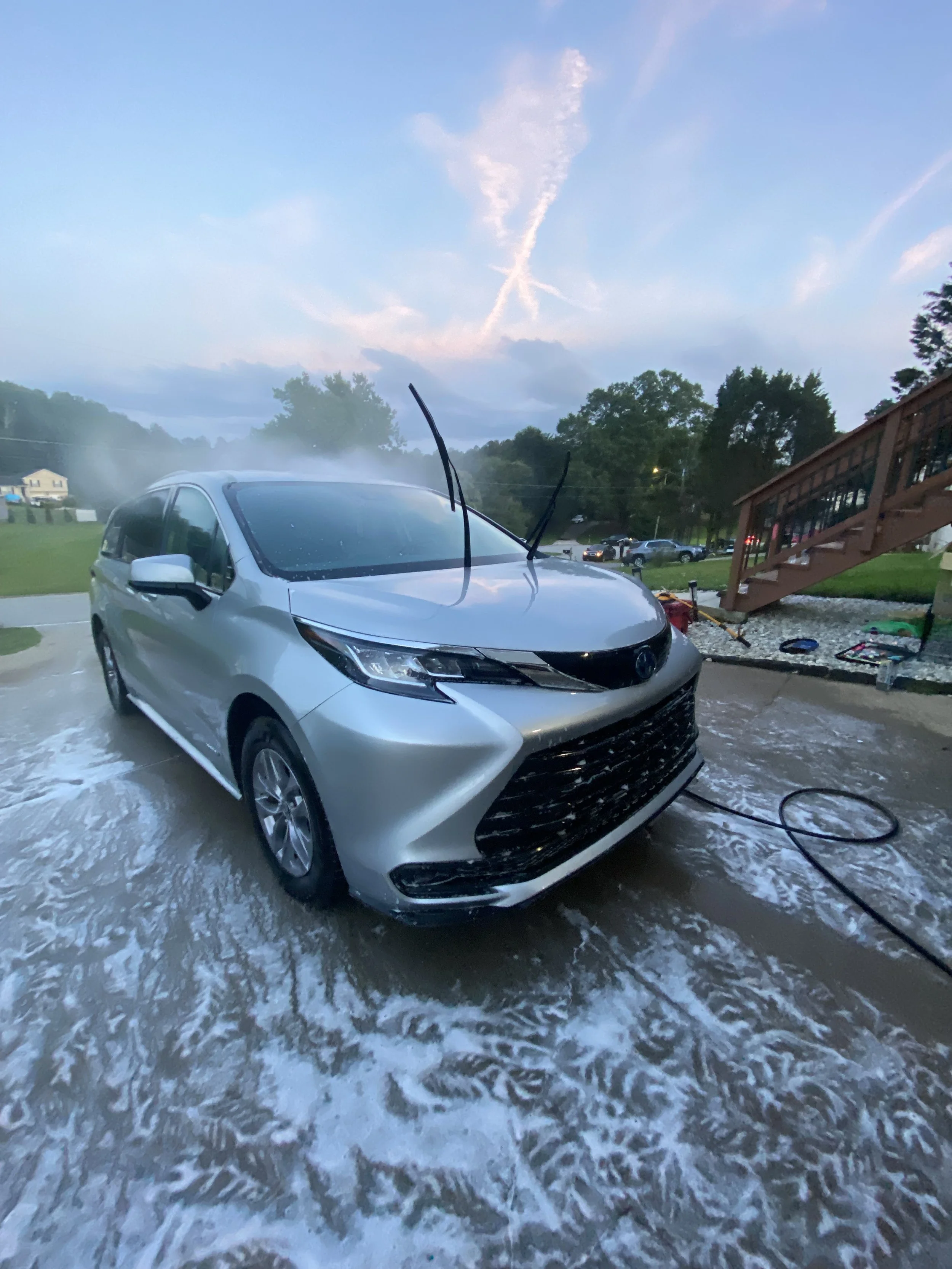 A silver Toyota minivan being washed with soap and water in a driveway during early evening, with a cloudy sky and some trees in the background.