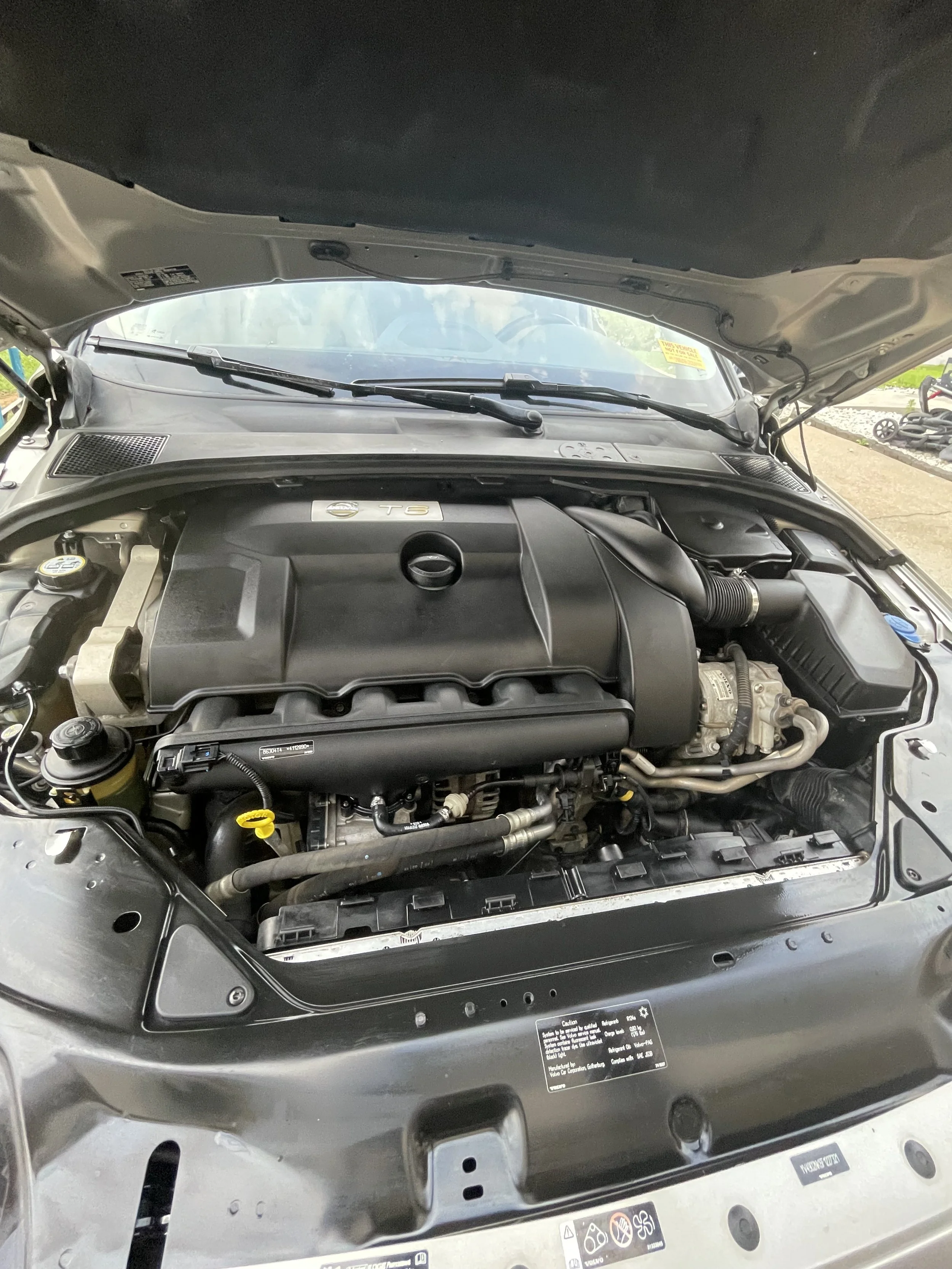 Engine bay of a car showing a black engine cover labeled T6, with various components and hoses visible.