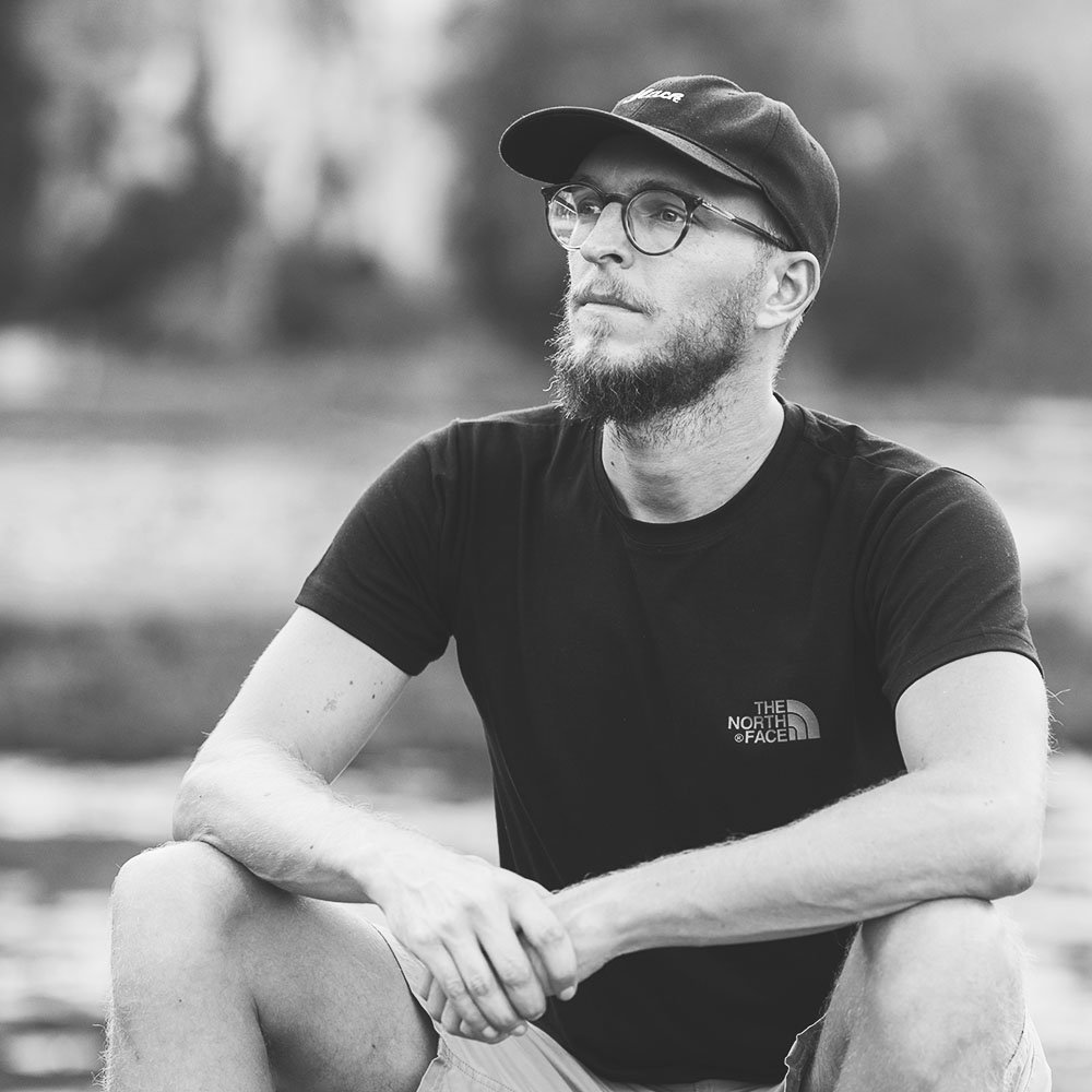 Schwarz-weiß-Foto eines Mannes mit Brille, Bärtchen, Basecap und T-Shirt von The North Face, sitzend im Freien, nachdenklich dreinblickend.