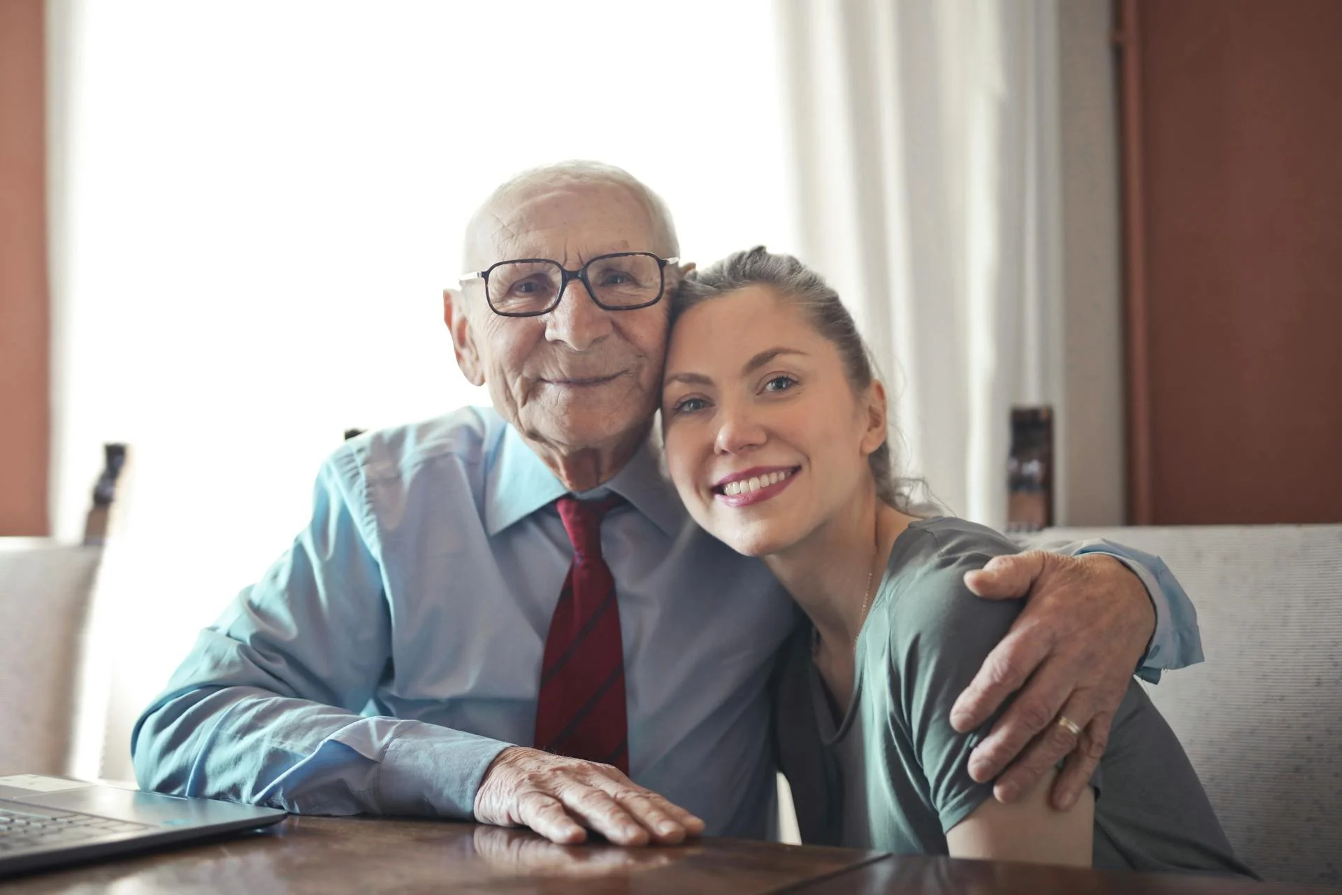An elderly man in glasses and a woman are sitting close together at a table, smiling and embracing each other, with sunlight streaming through a window behind them.