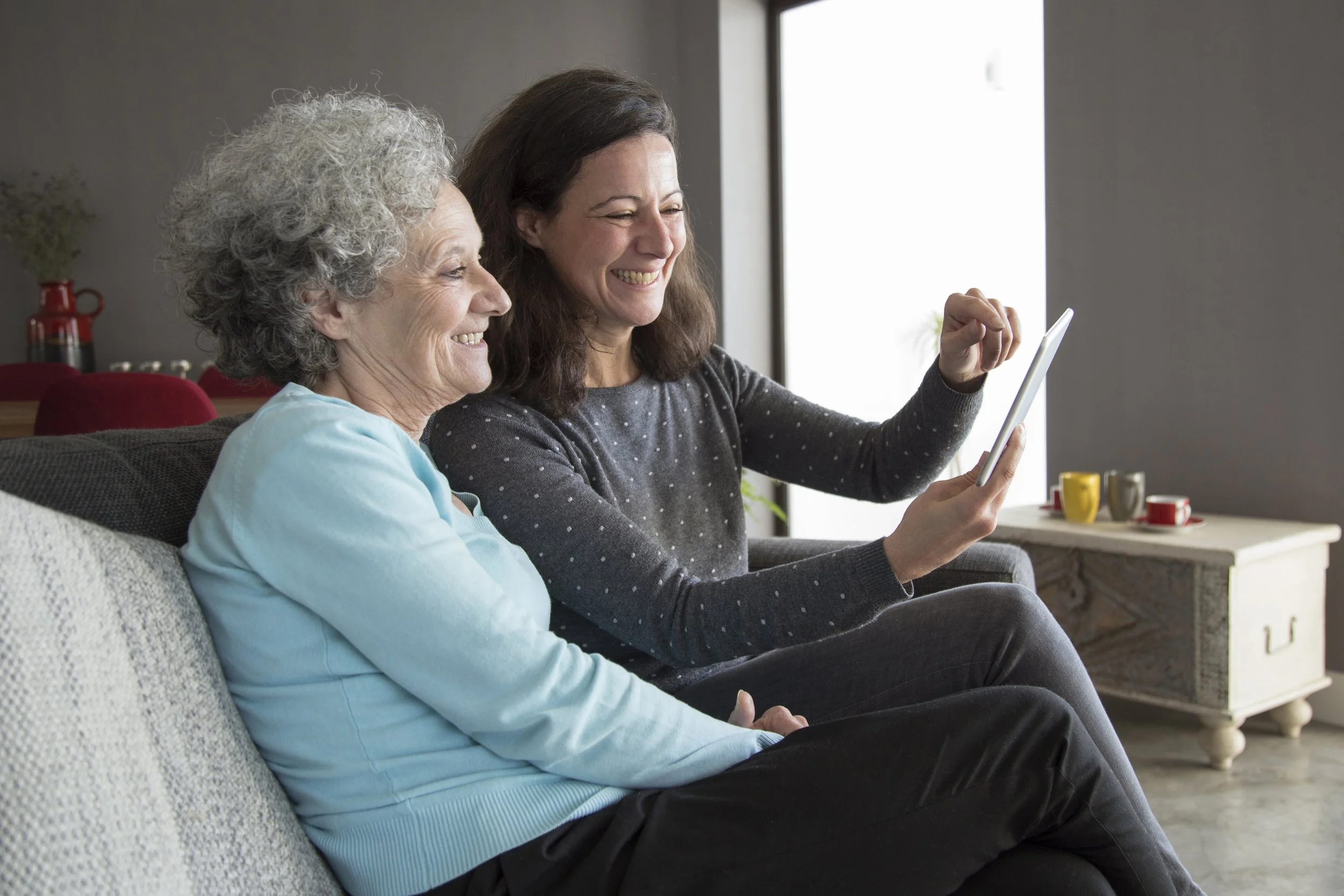 A senior woman and a middle-aged woman sitting on a sofa, smiling, looking at a tablet together in a cozy living room.