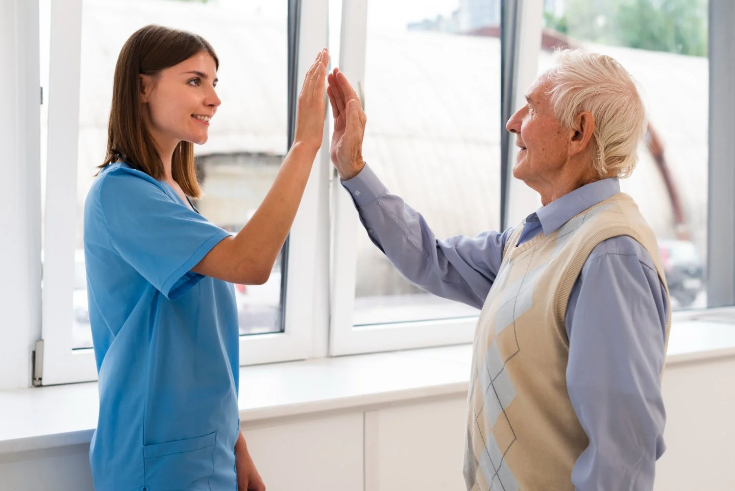 A young female healthcare worker giving an elderly male patient a high-five in a bright room with large windows.