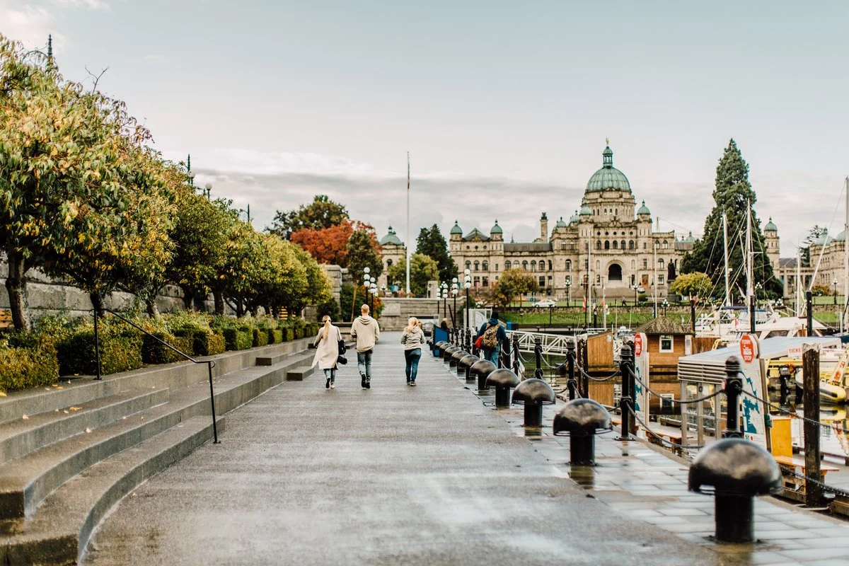 People walking along a waterfront in front of a large historic government building, with boats docked on the right and trees on the left.