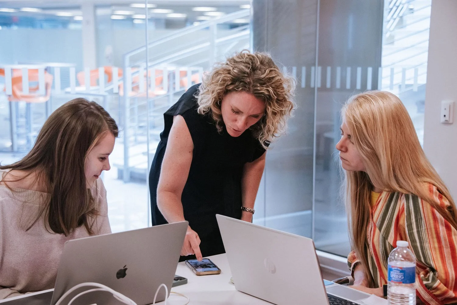 Three women in a meeting, two seated at laptops and one standing, all engaged in discussion in a bright office space.