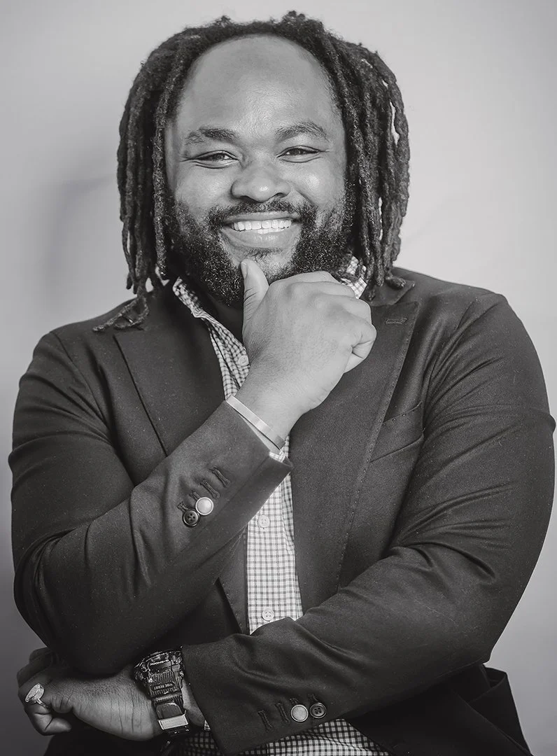 A smiling man with dreadlocks and a beard, wearing a suit jacket over a checkered shirt, with one hand on his chin and the other arm crossed.