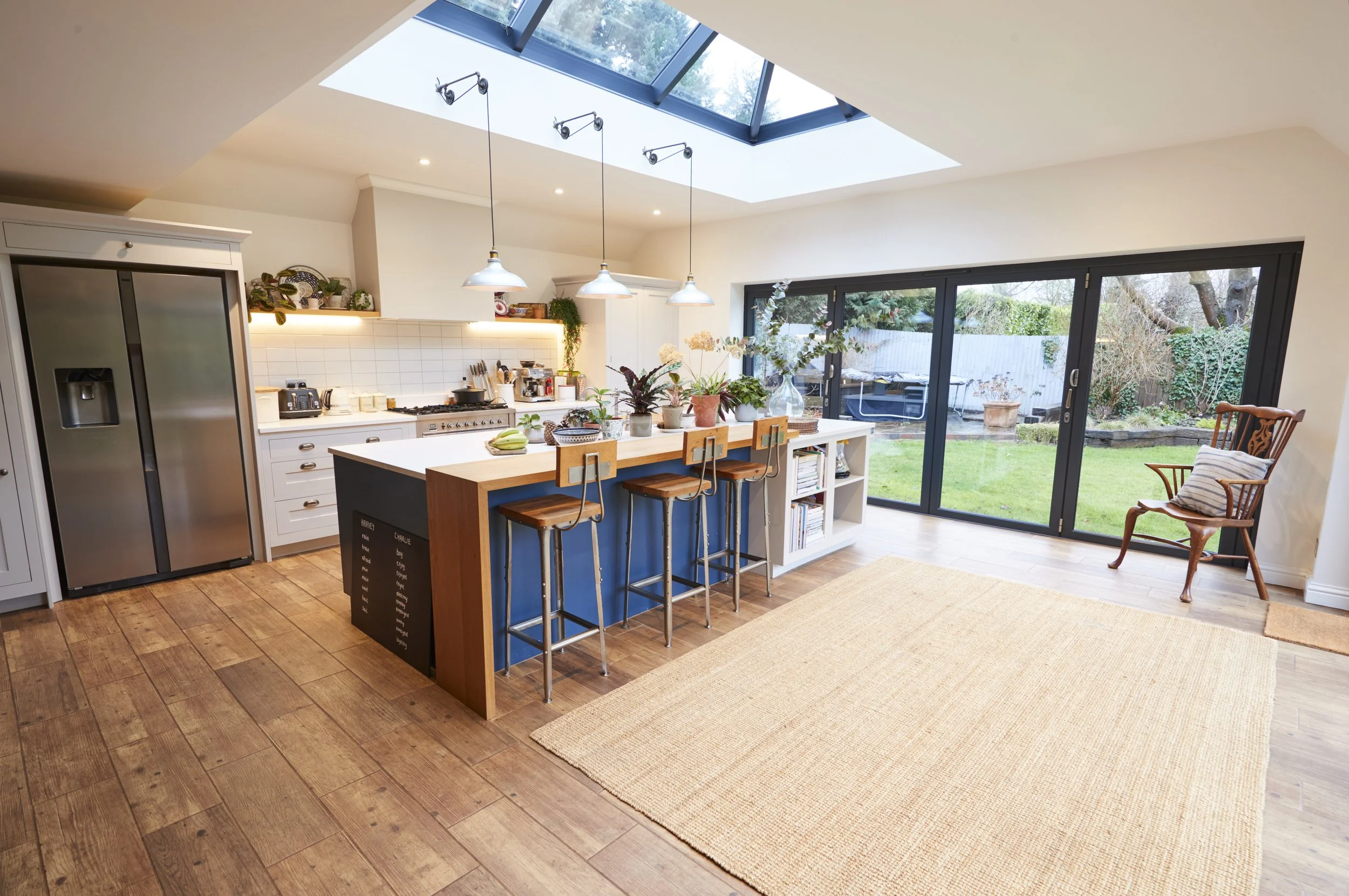 A modern kitchen with a skylight, wooden floor, white cabinetry, a large kitchen island with plants, stools, and a sliding glass door leading to a backyard.