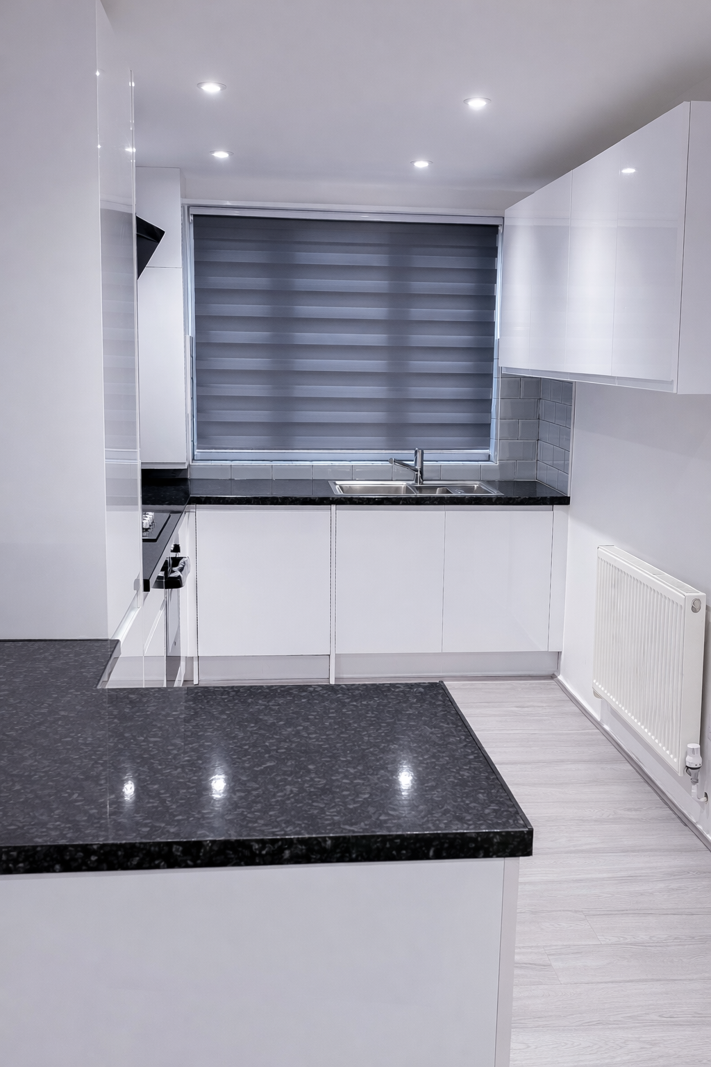 Modern kitchen with white cabinets, black granite countertops, stainless steel sink, gray window blinds, white subway tile backsplash, and built-in stovetop, with a white radiator on the right and recessed ceiling lights.