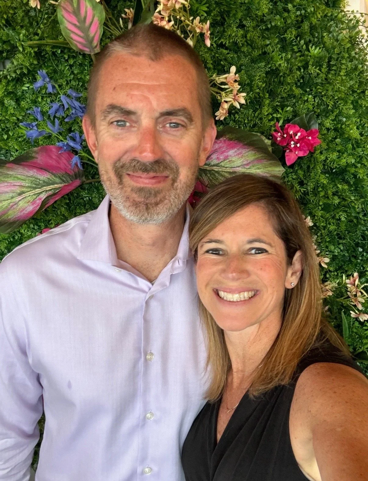 A smiling man and woman taking a selfie in front of a green leafy backdrop with colorful flowers.
