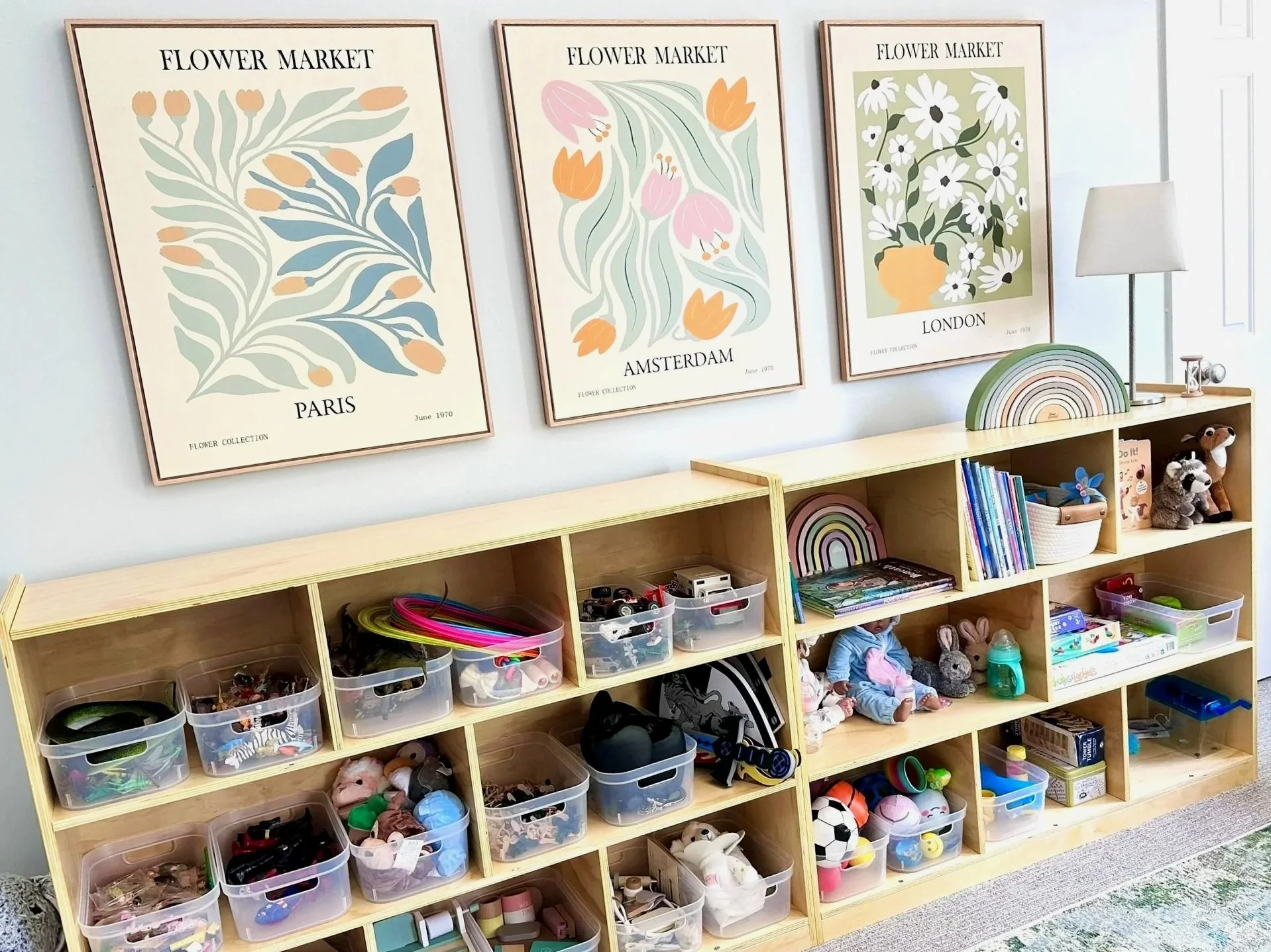 A wooden cubby shelving unit filled with bins of toys and stuffed animals, with several framed floral art prints on the wall above, titled 'Flower Market' from Paris, Amsterdam, and London, with simple, colorful floral illustrations.