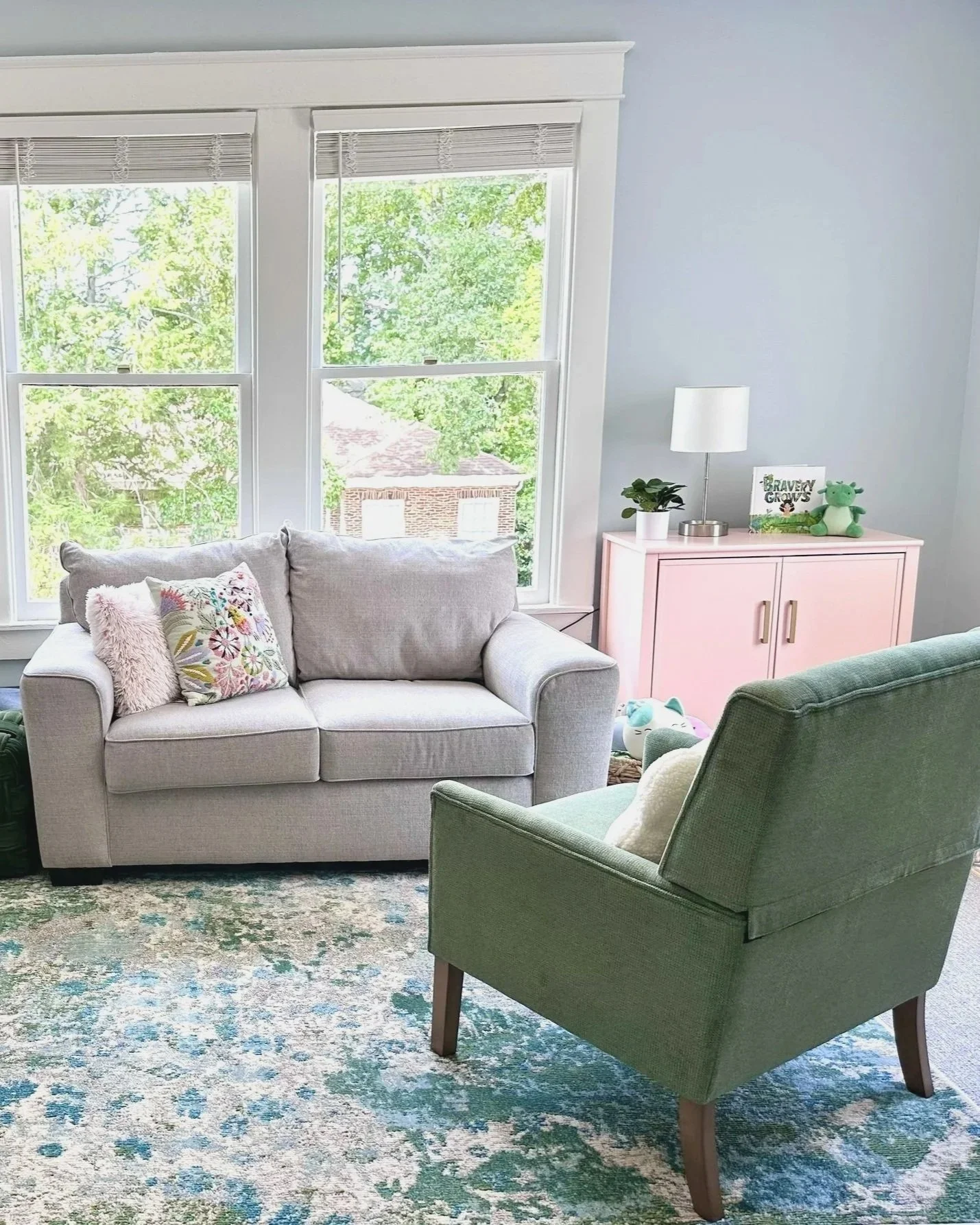 A cozy living room with a light gray sofa adorned with decorative pillows, facing a large window with a view of green trees and a brick building roof, a small pink cabinet with toys and a potted plant, a white table lamp, and a patterned area rug.
