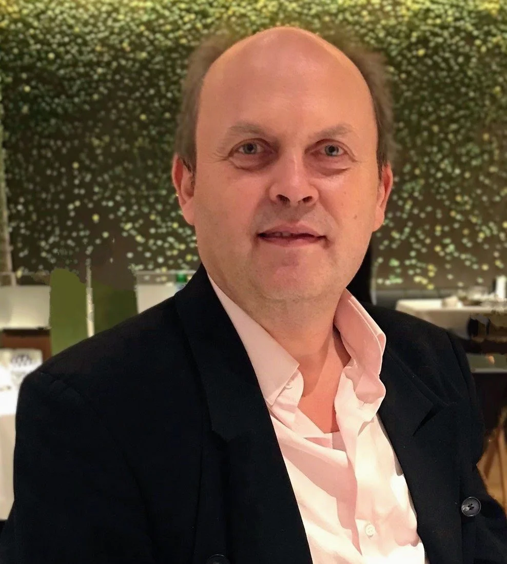 A middle-aged man with short hair, wearing a black blazer and a light pink shirt, sitting in a restaurant with a decorative backdrop of illuminated branches behind him.
