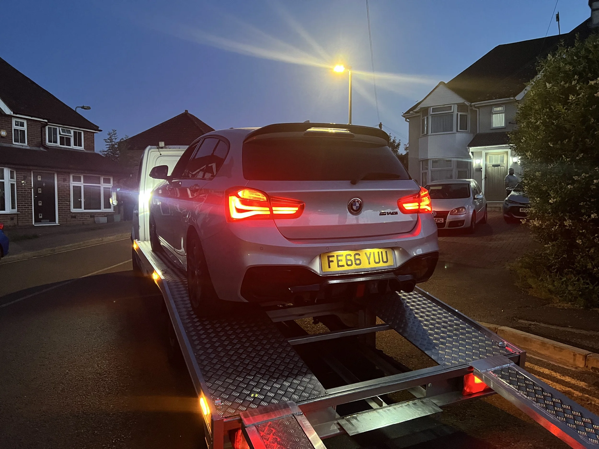 A silver BMW vehicle is on a flatbed tow truck at twilight in a residential neighborhood with houses and other parked cars nearby.