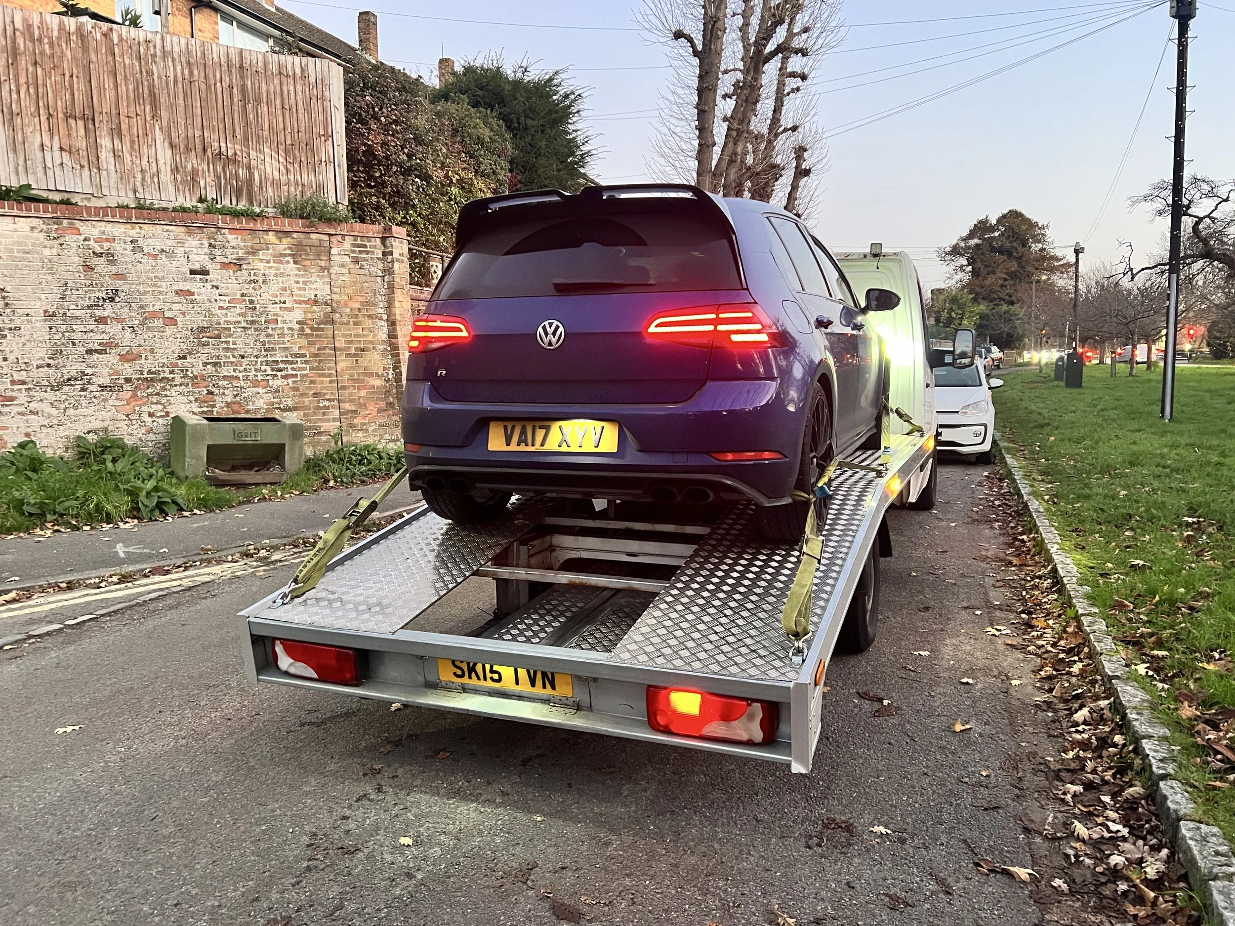 A purple Volkswagen golf hatchback loaded onto a flatbed tow truck parked on the side of a residential street. The license plates are visible, and the tow truck is equipped with yellow straps securing the car.