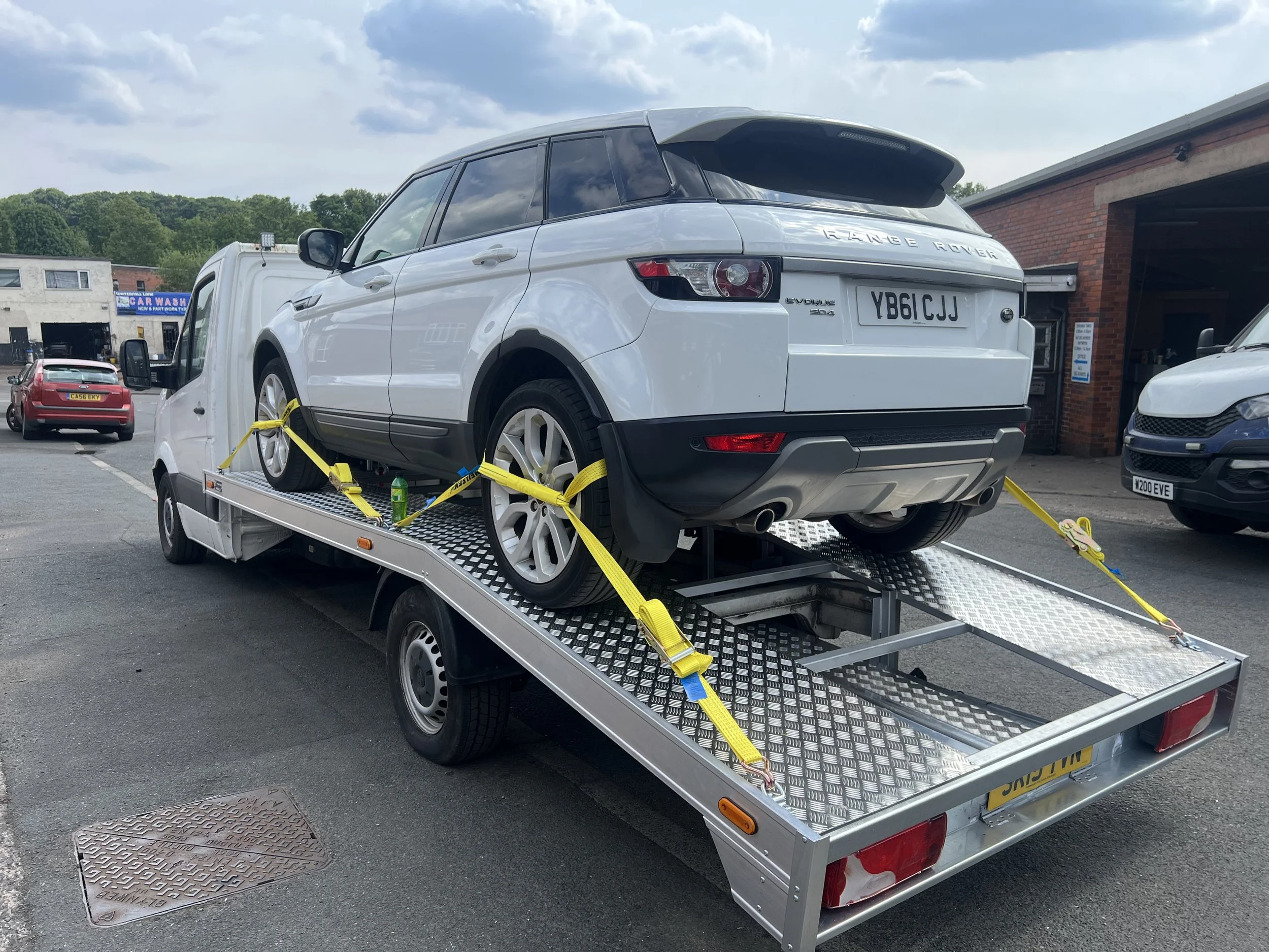 A white Range Rover Evoque secured on a flatbed tow truck with yellow straps, in a parking lot near a car wash, with several other vehicles visible in the background.