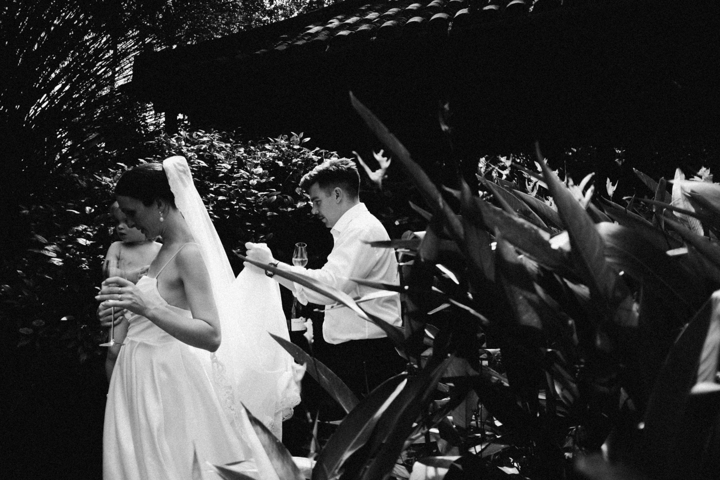 A black and white photo of a bride and groom at an outdoor wedding, with the bride holding a drink and the groom looking at something in his hand, surrounded by lush foliage.