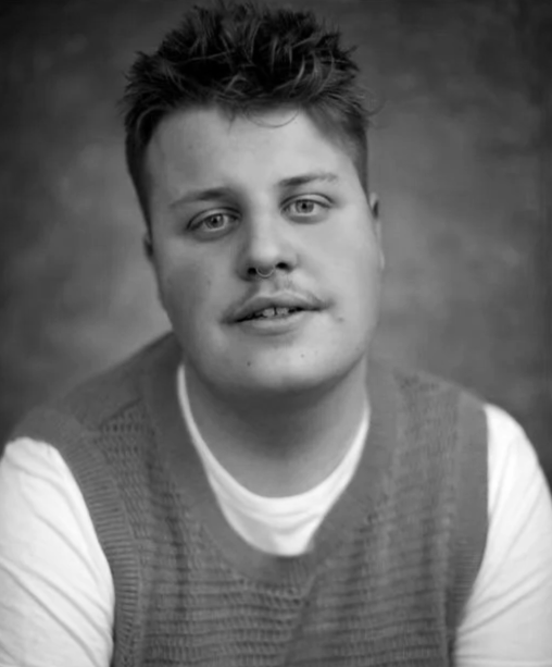 Black and white portrait of a young man with short, tousled hair, wearing a light-colored T-shirt and a knitted vest, looking at the camera with a neutral expression.