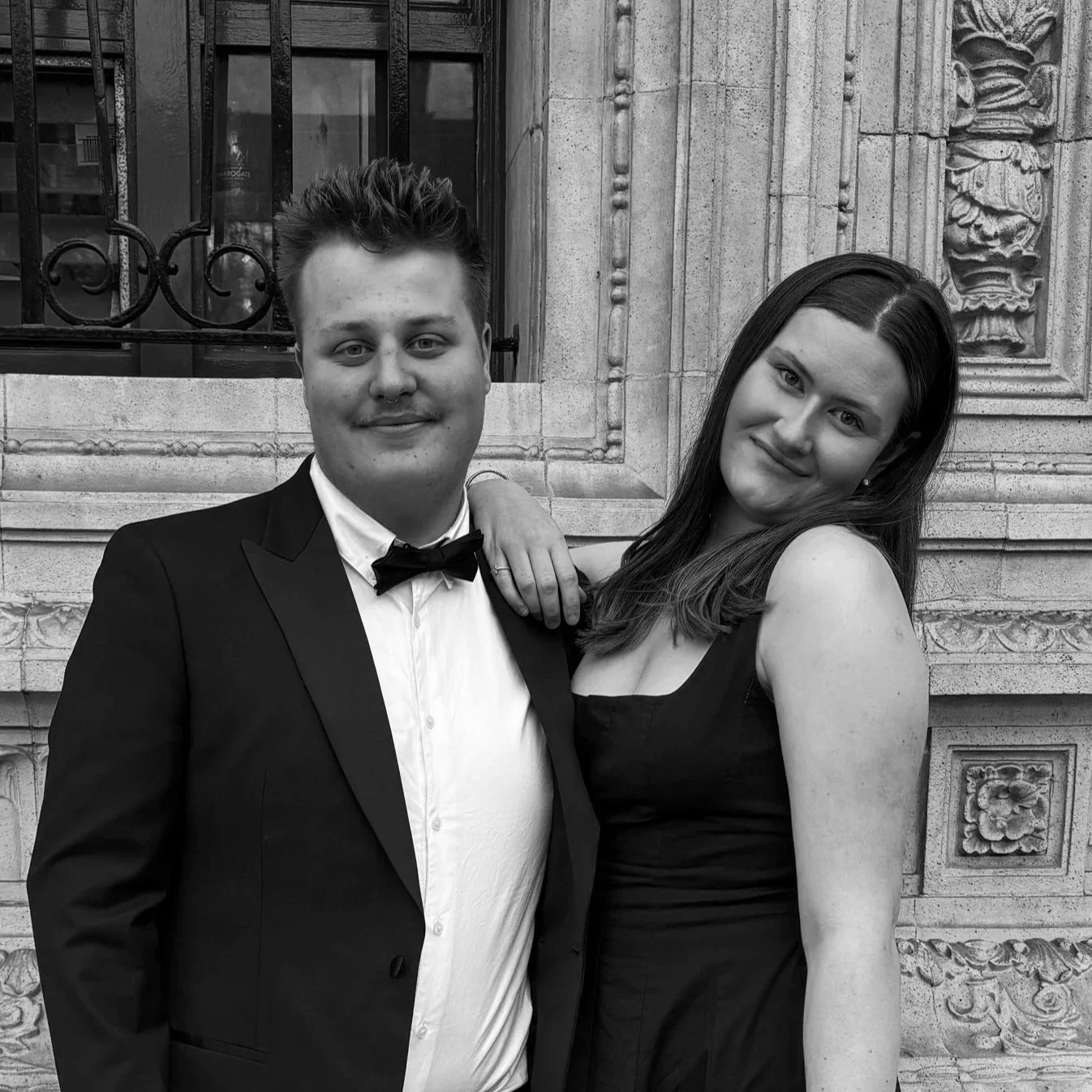 A young man and woman in formal attire standing in front of an ornate stone building, smiling at the camera.