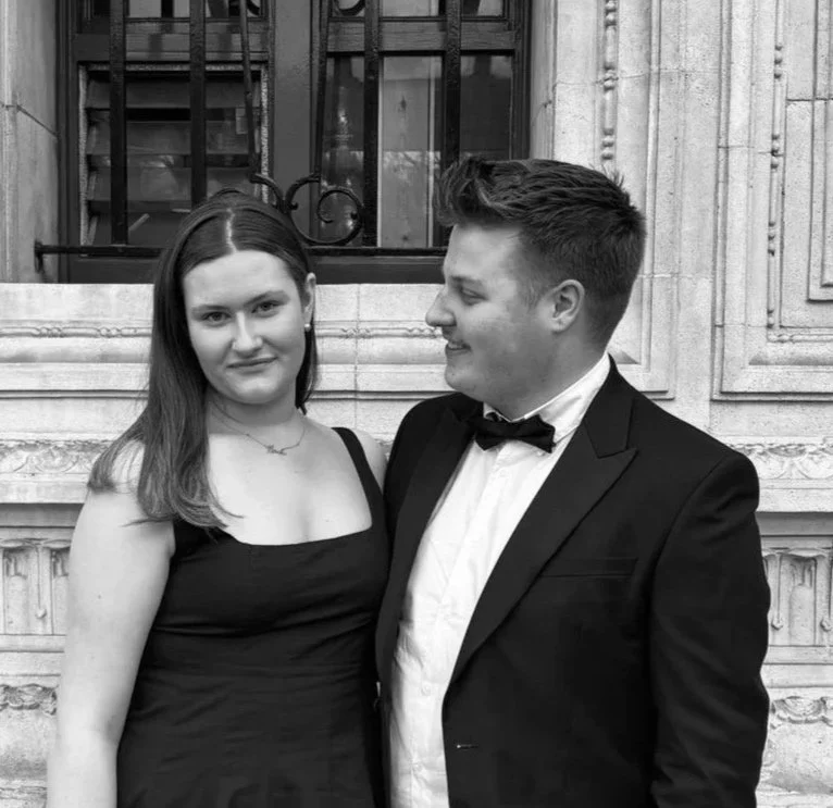 A young woman in a black dress and a young man in a tuxedo are standing closely together in front of a building with ornate architectural details and a window with iron bars.