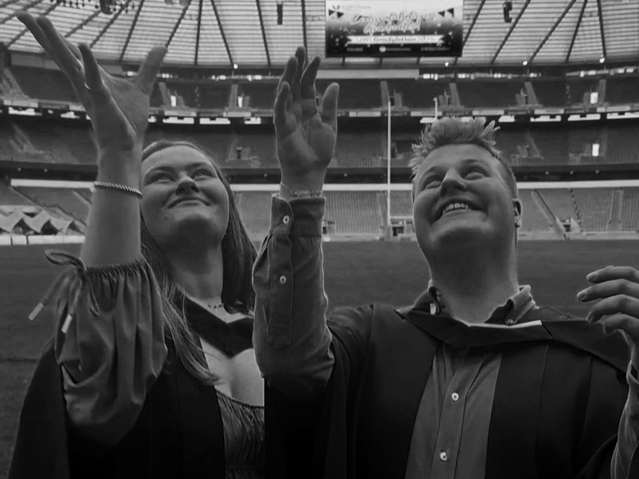 Two graduates in caps and gowns celebrating on a football field, with a stadium in the background.