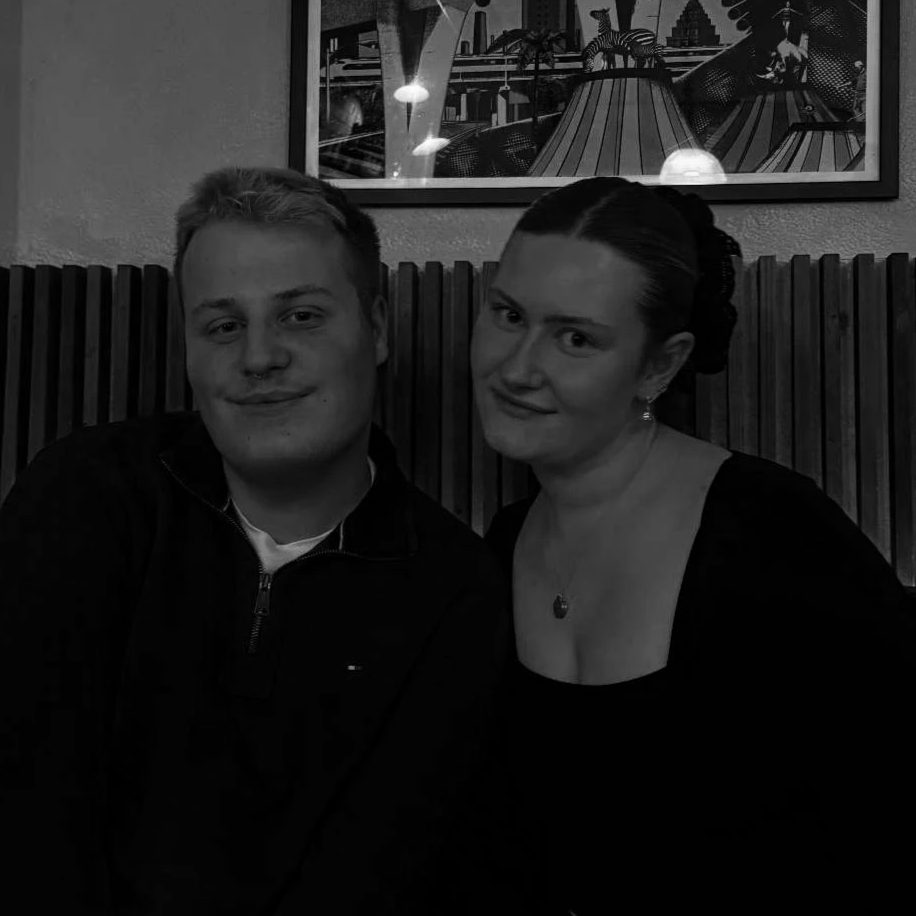 A man and a woman sitting close together in a restaurant, posing for the camera with smiles. The background features a poster or picture of a futuristic cityscape.