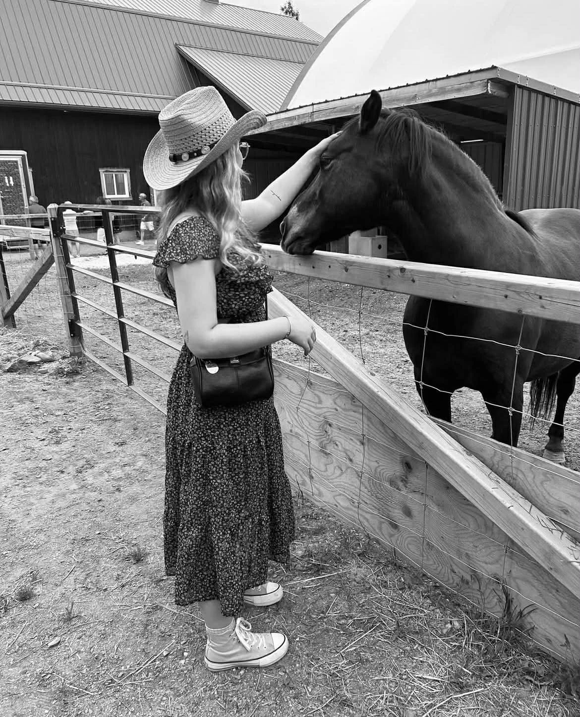 Une jeune femme portant un chapeau de paille, une robe à fleurs, des baskets blanches et un sac noir, caresse la tête d'un cheval dans un enclos.