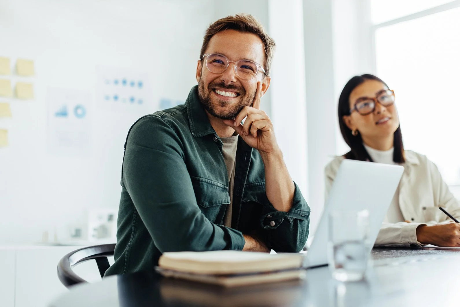 Hombre sonriendo en una reunión de oficina, con una mujer al fondo tomando notas.