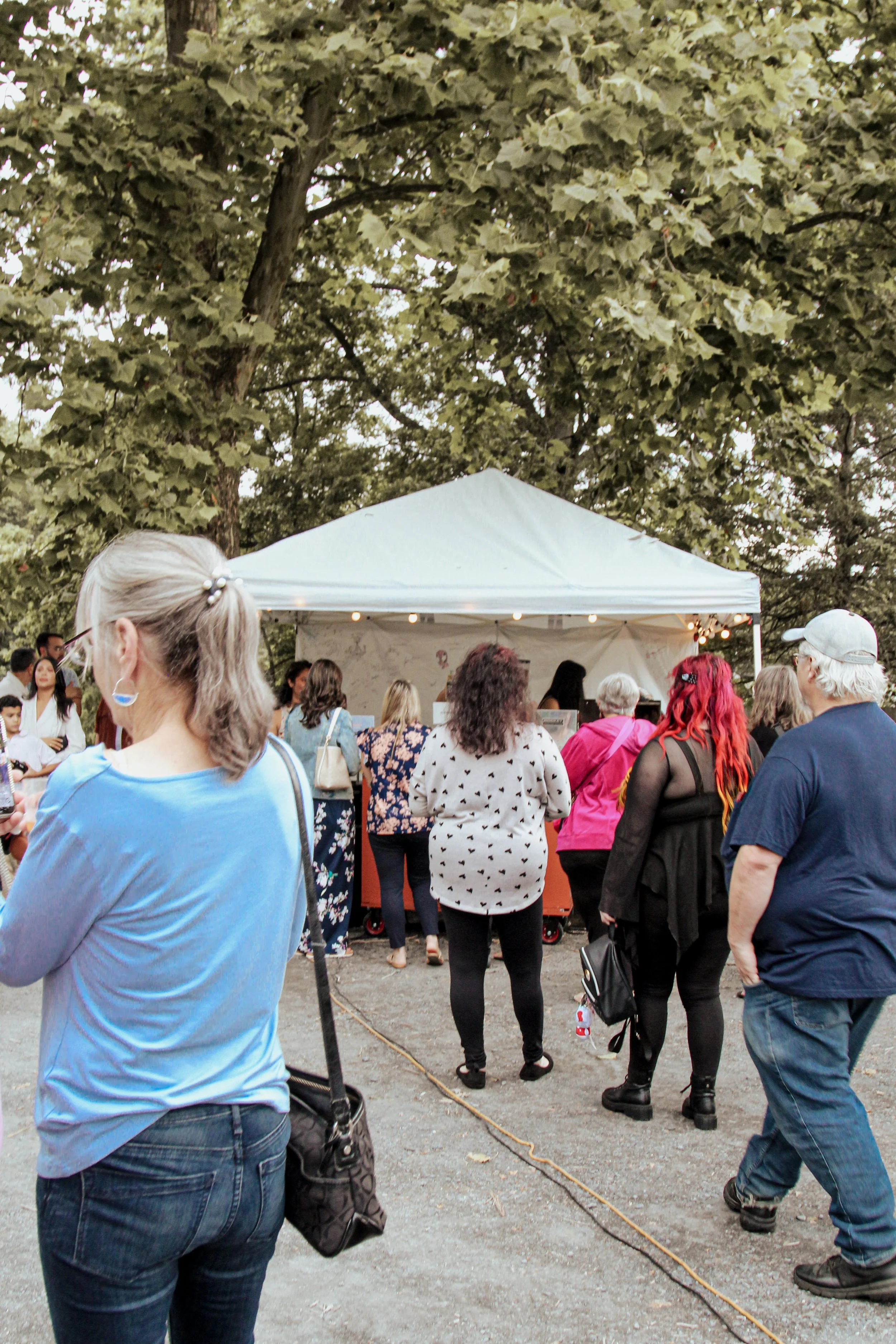 A group of people gathered around a white canopy tent outdoors, under large trees, at what appears to be a community event or festival.