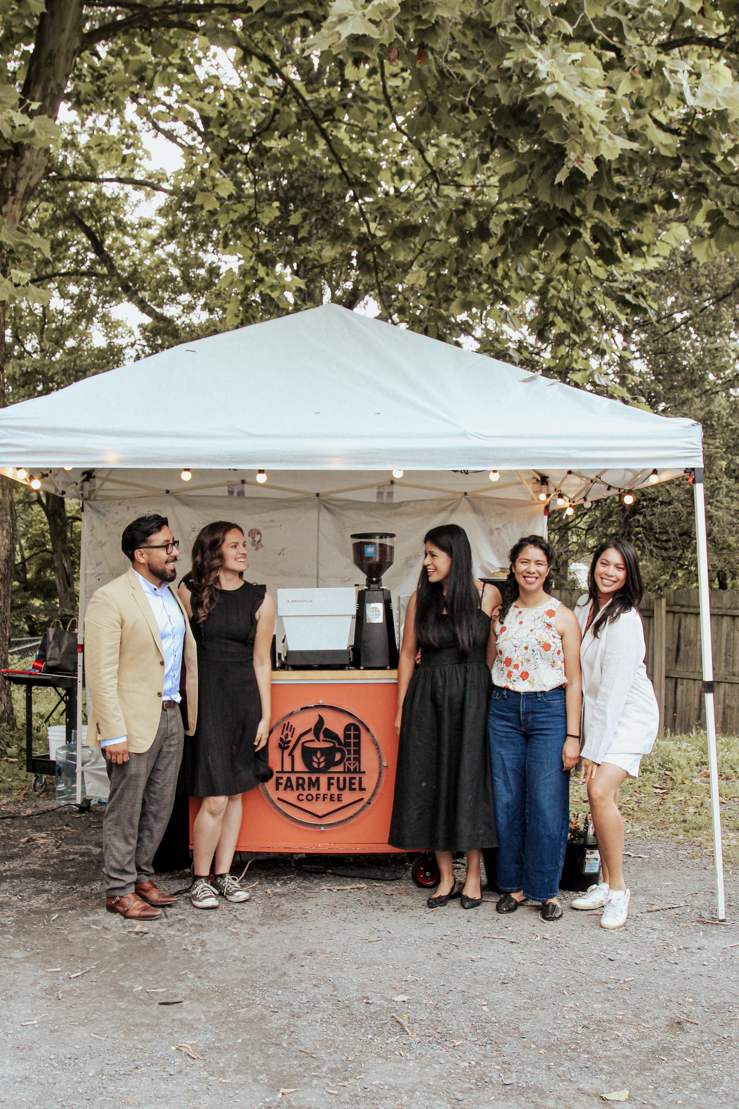 Group of five people standing in front of a coffee stand labeled 'Farm Fuel Coffee,' under a white canopy, outdoors in a wooded area.