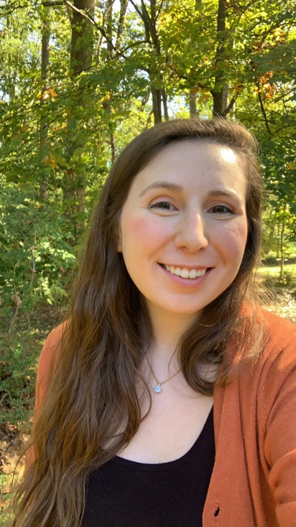 A smiling woman with long brown hair taking a selfie outdoors in a wooded area with green leaves.