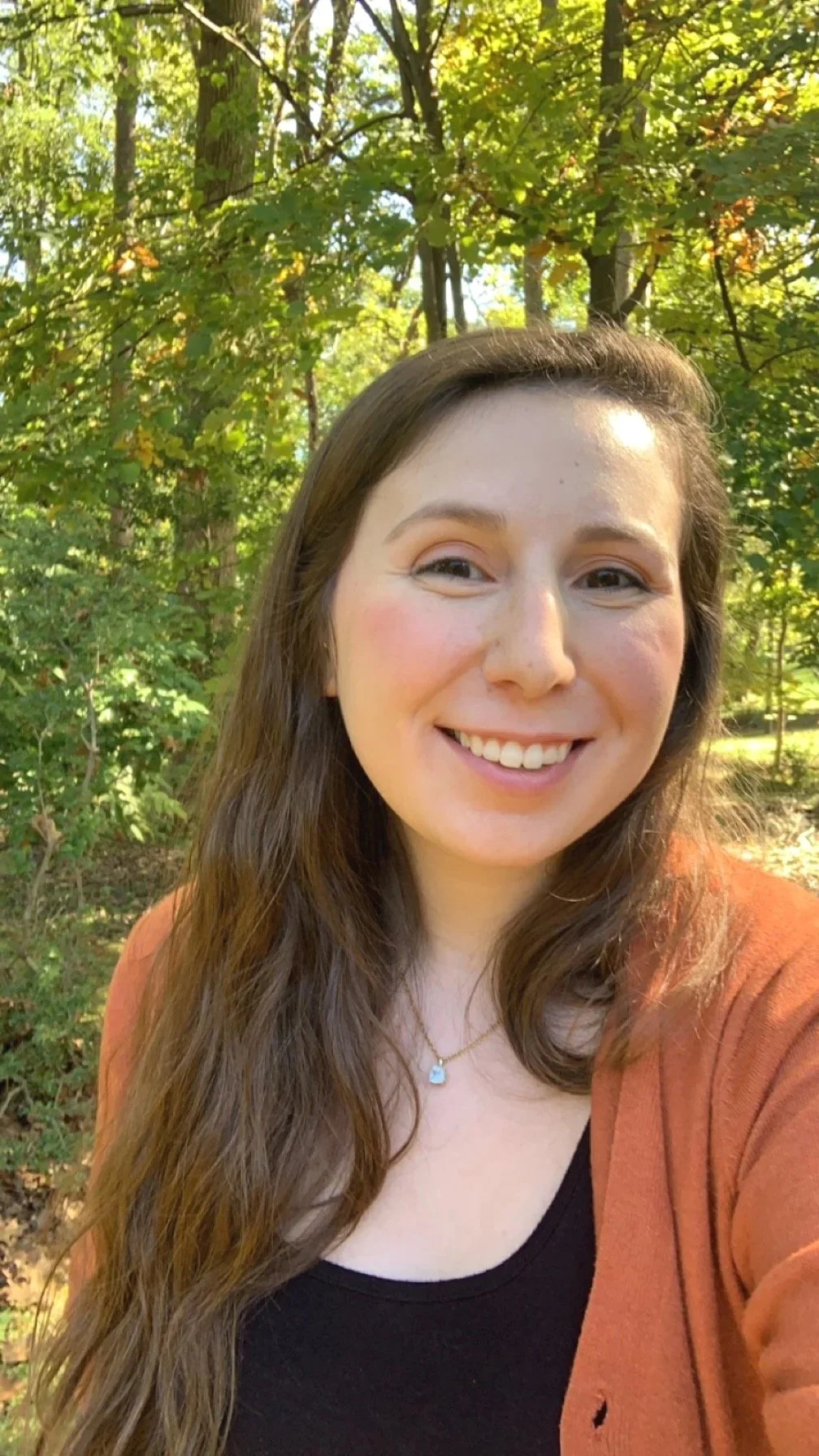 A woman with long brown hair smiling outdoors with green trees and sunlight in the background.