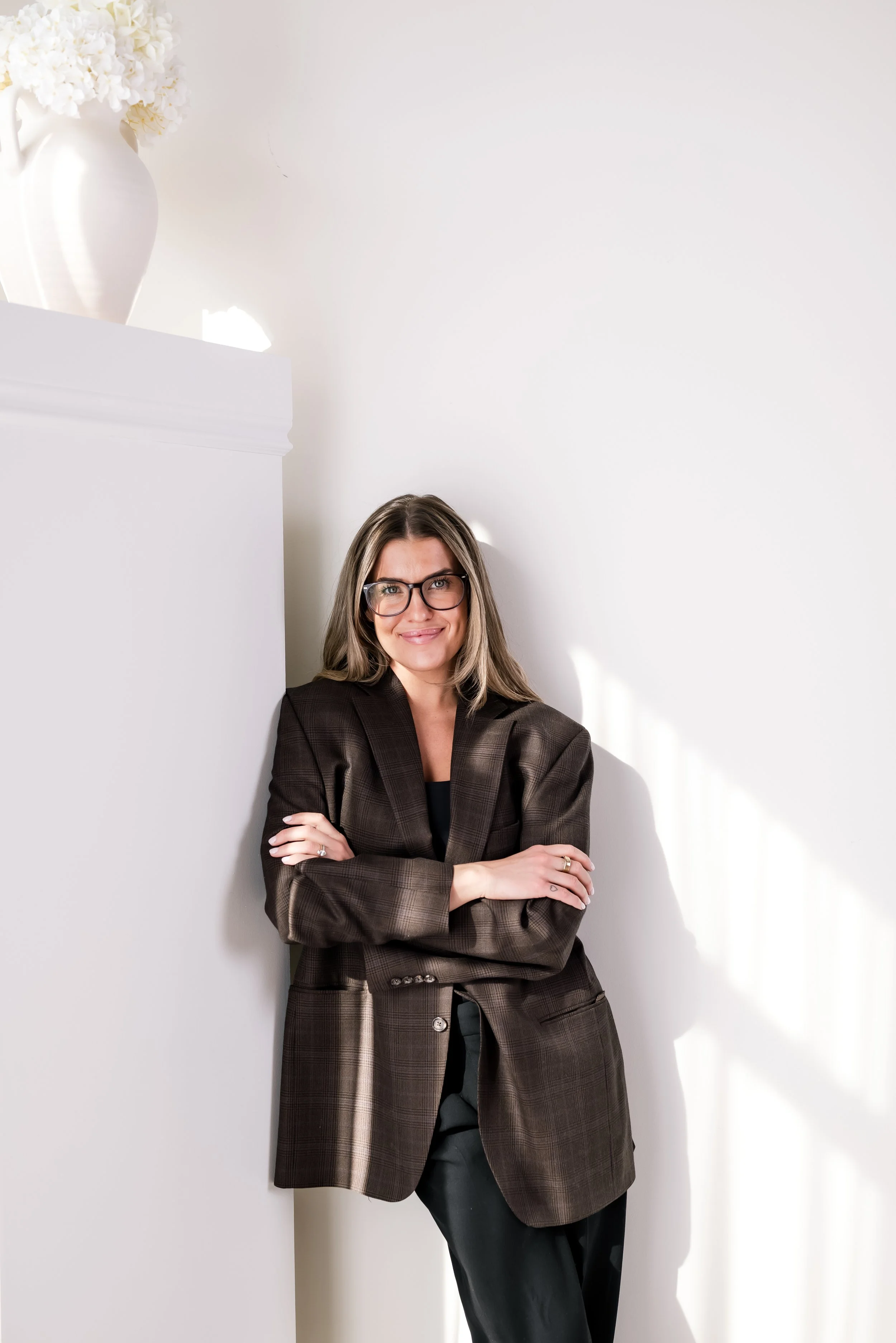 A woman with shoulder-length hair, glasses, wearing a brown plaid blazer, standing against a white wall and leaning with her arms crossed.