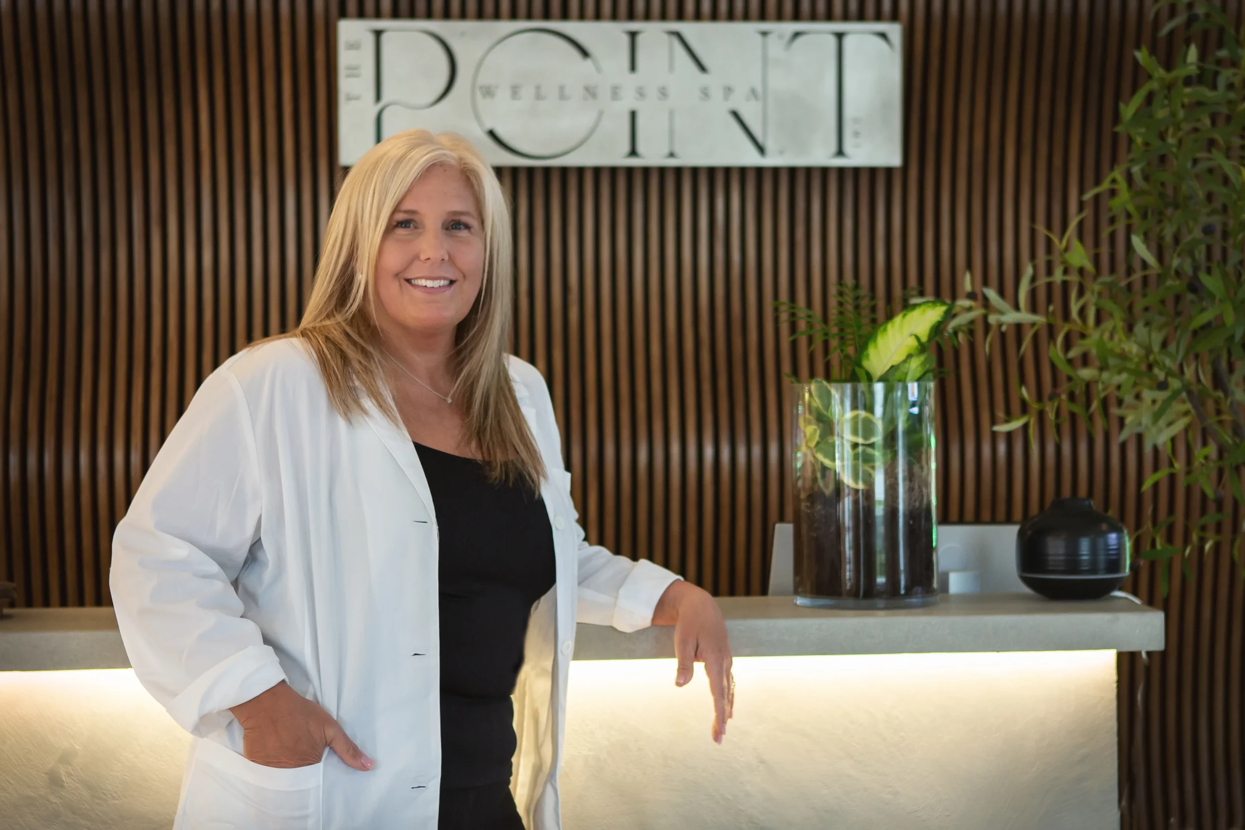 A woman with blonde hair smiling, wearing a white coat and black top, standing in front of a wellness spa reception with a wooden wall, a sign that reads 'THE POINT WELLNESS SPA,' a glass vase with green leaves, and a black decorative object.