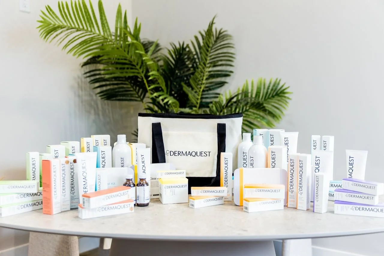 Skincare products arranged on a table with a white tote bag marked 'DERMAQUEST' in front of a green plant.