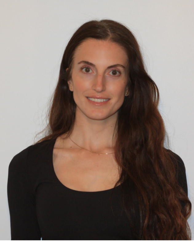 A woman with long, wavy brown hair wearing a black top stands against a plain white background, smiling at the camera.